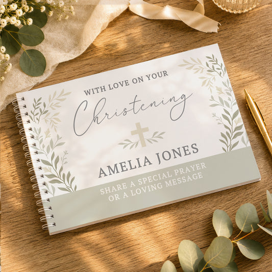A personalised spiral-bound Christening guest book with a white cover, sage green botanical border illustrations, and a gold cross, displayed on a wooden tabletop with eucalyptus leaves, a gold pen, and a cream ribbon.