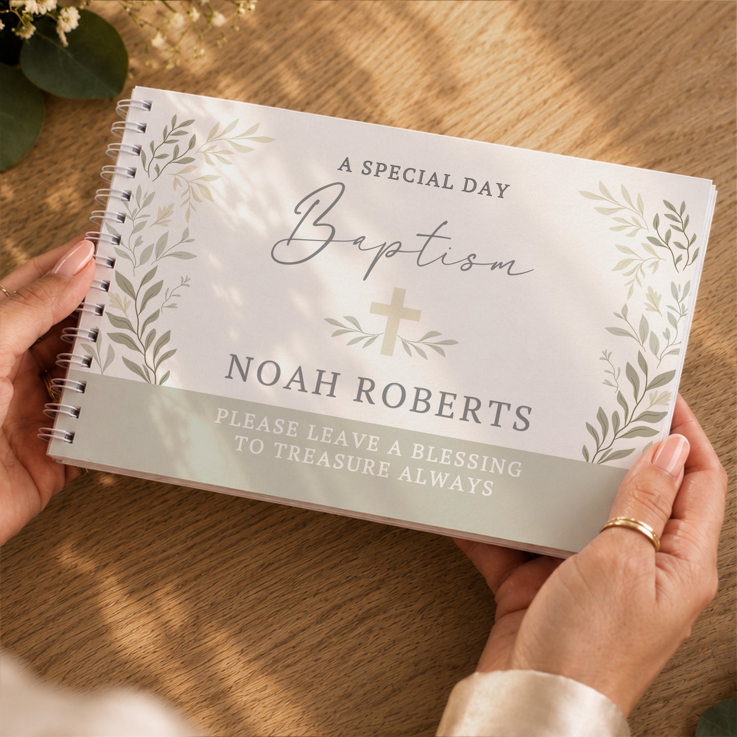 A personalised spiral-bound baptism keepsake guest book held by two hands over a wooden table. The white cover features elegant grey script reading 'Baptism', a small gold cross, and floral illustrations, with the name 'NOAH ROBERTS' printed in grey.