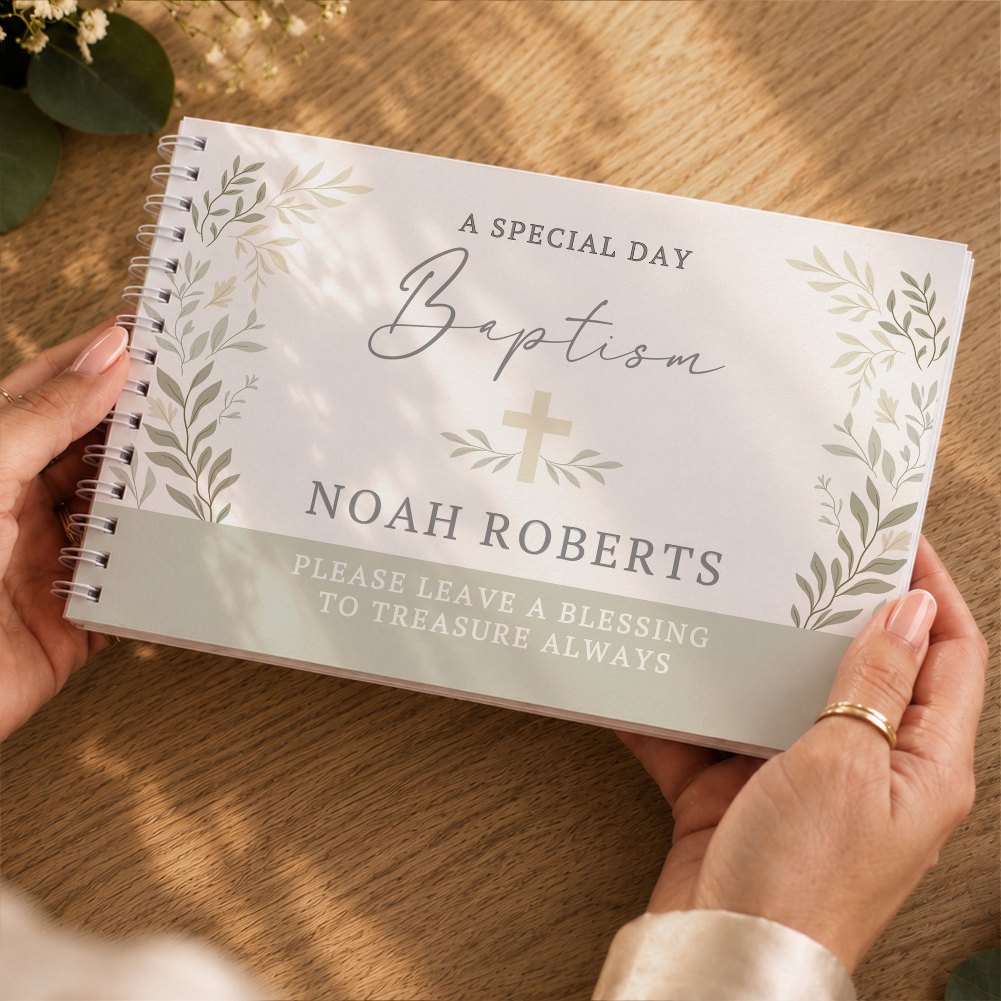 A personalised spiral-bound baptism keepsake guest book held by two hands over a wooden table. The white cover features elegant grey script reading 'Baptism', a small gold cross, and floral illustrations, with the name 'NOAH ROBERTS' printed in grey.