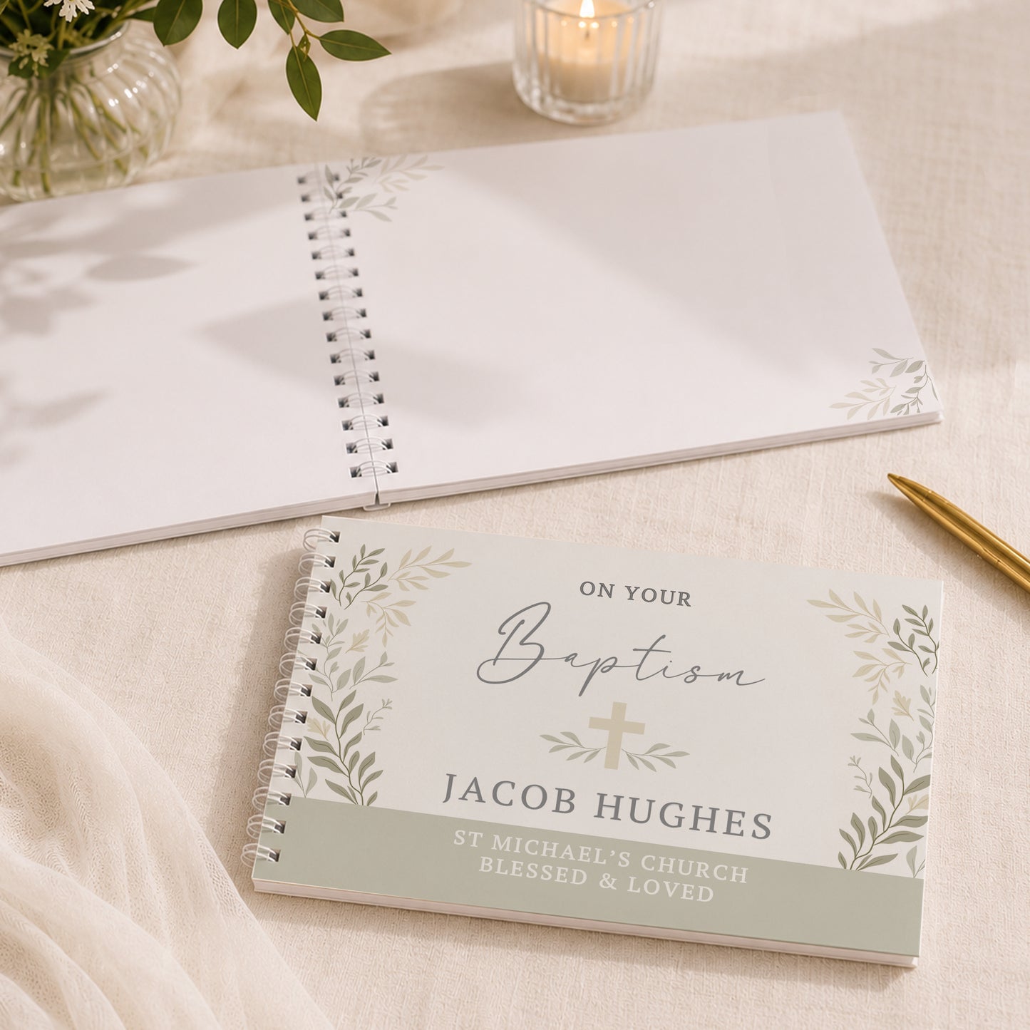 A personalised baptism guest book with a white cover, silver spiral binding, and green botanical illustrations, placed on a beige fabric surface next to a gold pen. Another blank guest book and a lit candle are visible in the background.