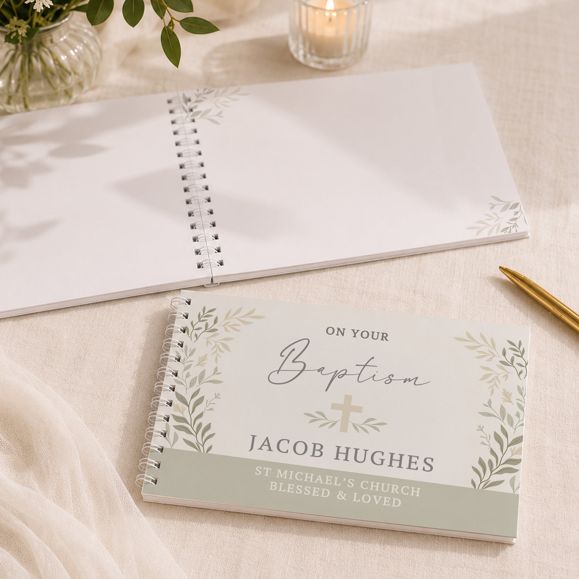 A personalised baptism guest book with a white cover, silver spiral binding, and green botanical illustrations, placed on a beige fabric surface next to a gold pen. Another blank guest book and a lit candle are visible in the background.