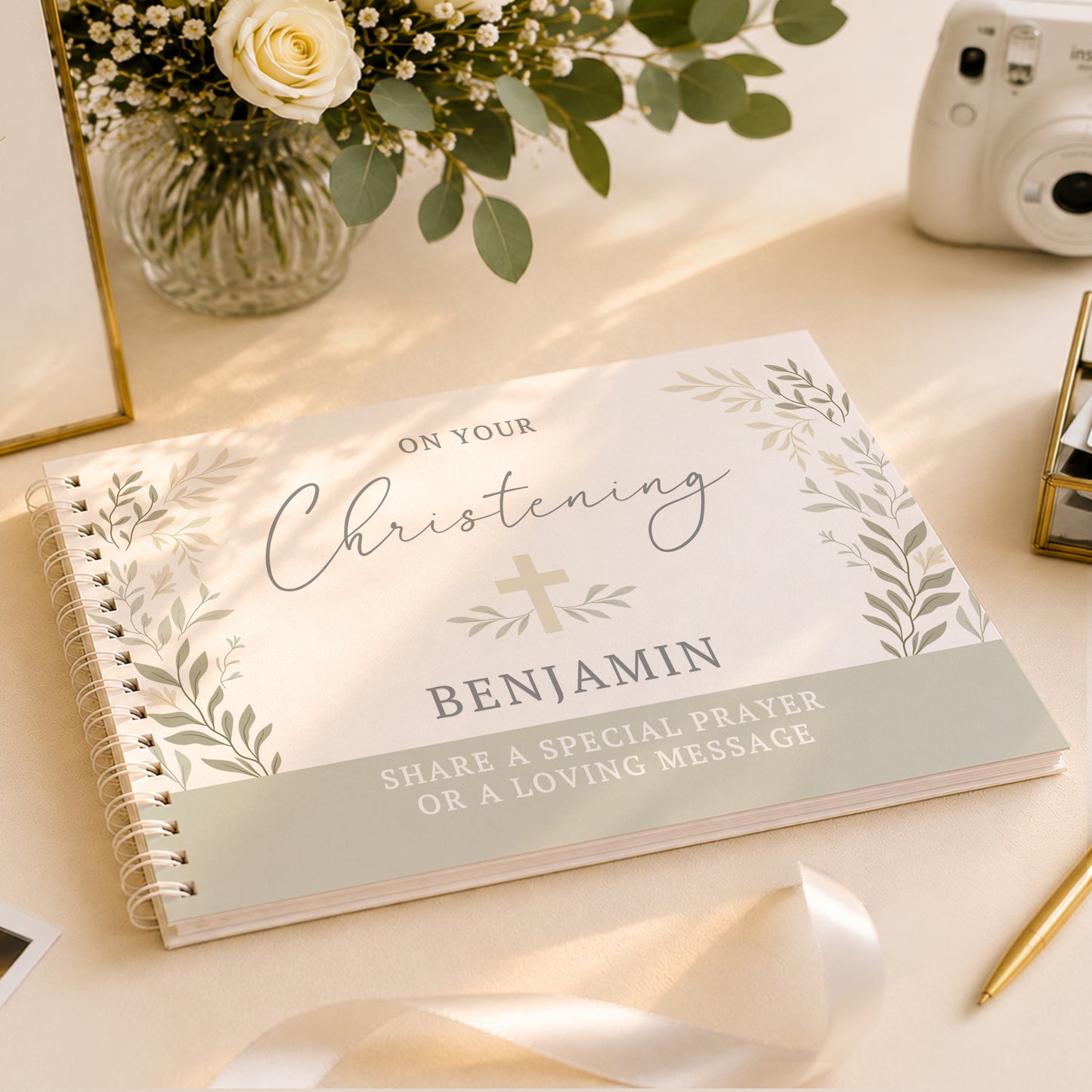 A personalised spiral-bound christening journal or gift book lies open on a light beige surface. The cover reads 'ON YOUR Christening BENJAMIN' with a small cross and leafy border, plus the message 'SHARE A SPECIAL PRAYER OR A LOVING MESSAGE.