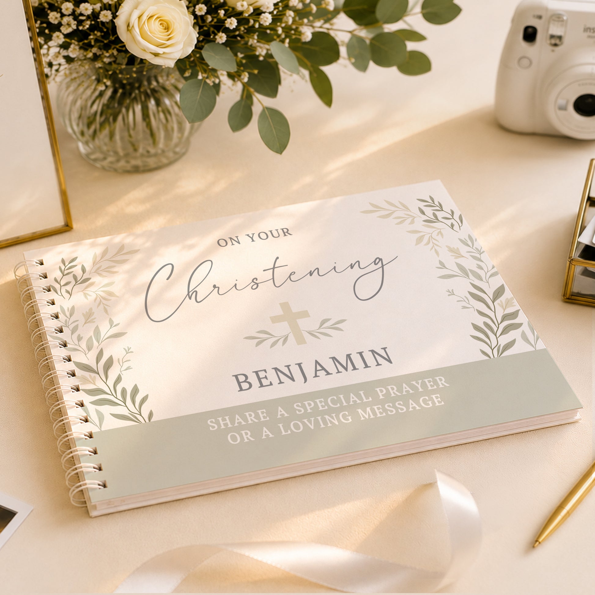 A personalised spiral-bound christening journal or gift book lies open on a light beige surface. The cover reads 'ON YOUR Christening BENJAMIN' with a small cross and leafy border, plus the message 'SHARE A SPECIAL PRAYER OR A LOVING MESSAGE.