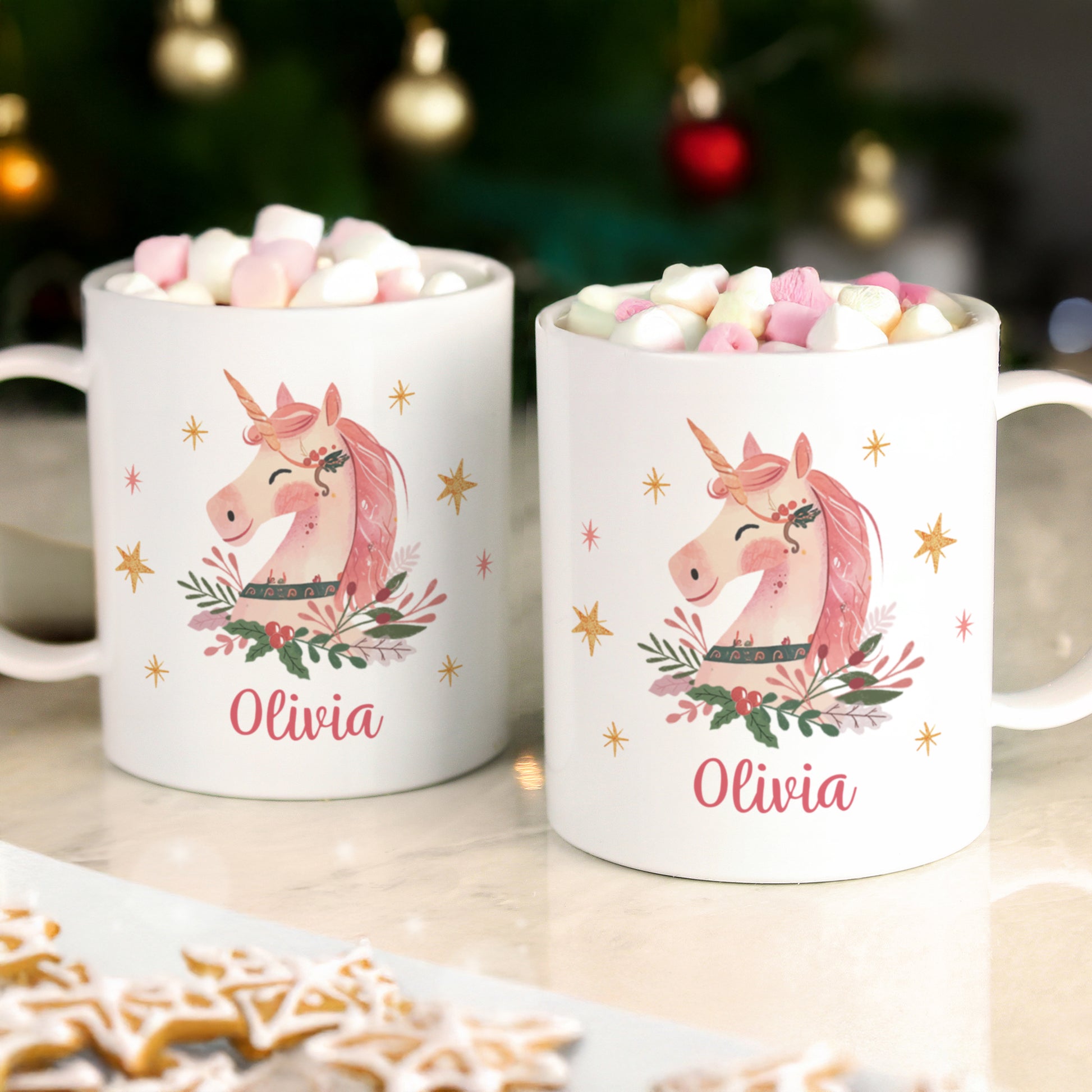 Two white mugs with unicorn designs and 'Olivia' text, filled with hot chocolate and marshmallows, on a table with a blurred Christmas tree background.