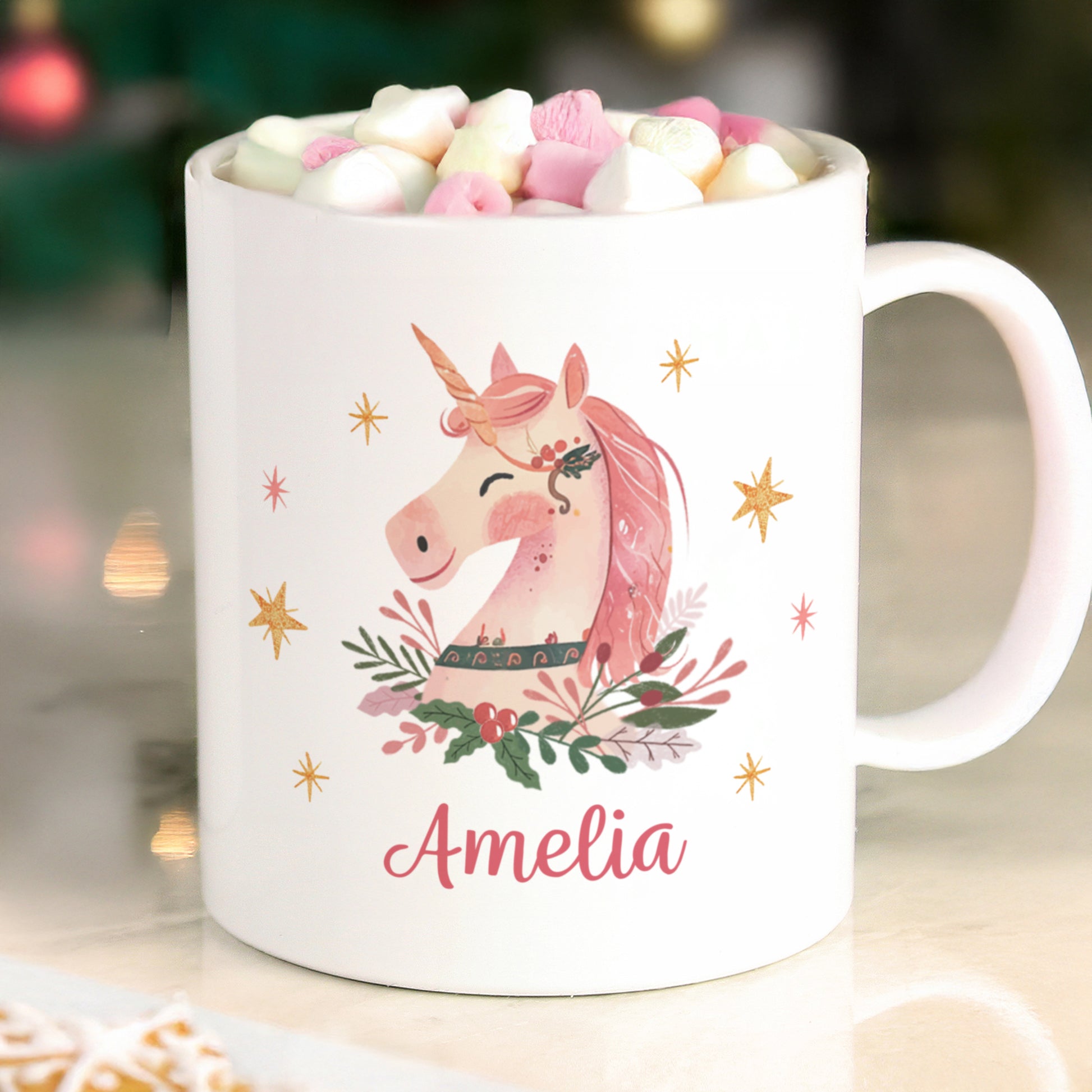 White mug with a pink unicorn design and the name 'Amelia', filled with hot chocolate and marshmallows, on a blurred background.