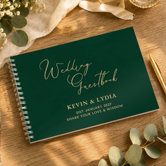A personalised wedding guestbook with a dark green cover and gold foil lettering, styled on a wooden table with eucalyptus leaves, a gold pen, and a cream ribbon.
