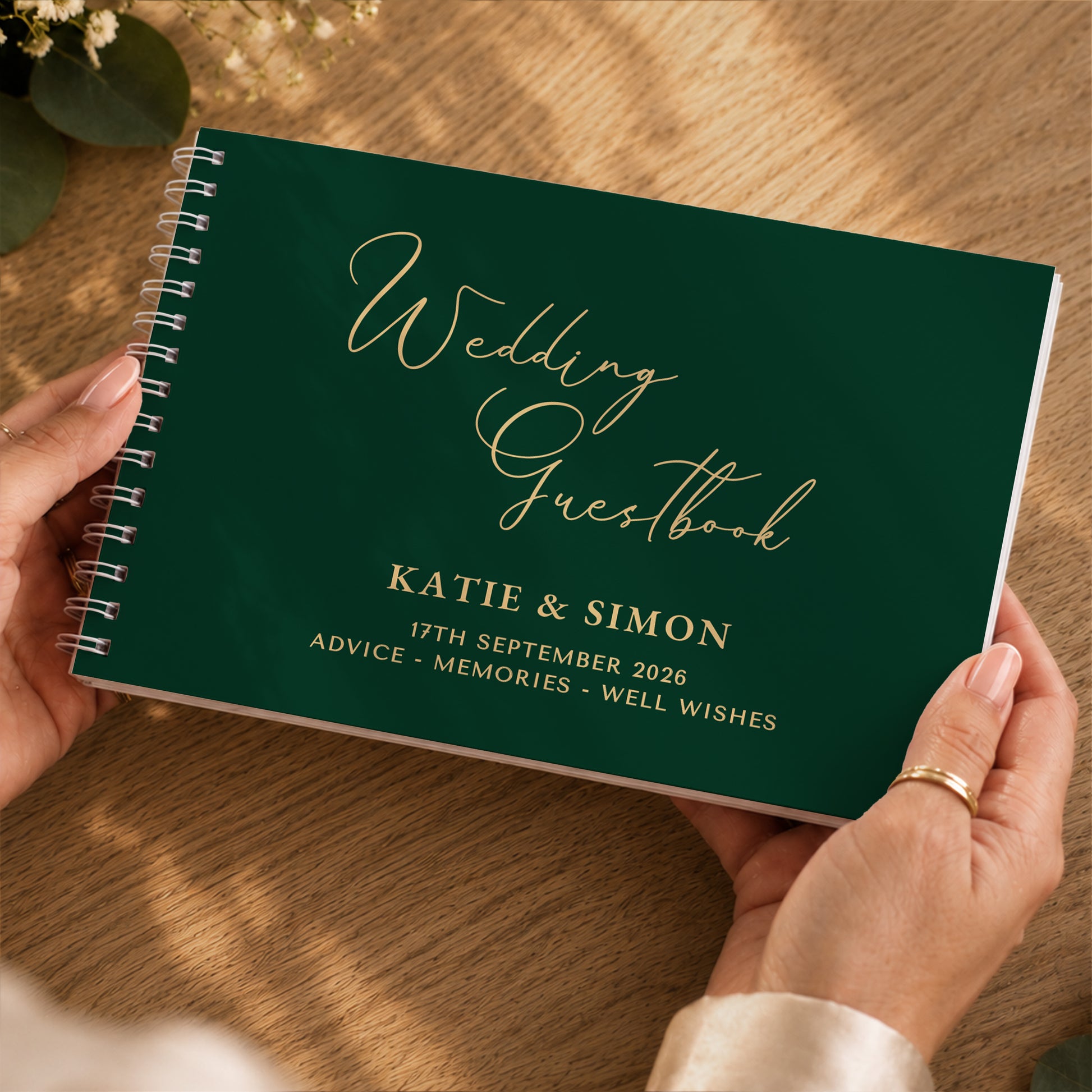 A dark green spiral-bound wedding guestbook being held by two hands over a wooden table. The cover features elegant gold script reading 'Wedding Guestbook' and gold printed details for 'Katie & Simon' with the date '17th September 2026'.