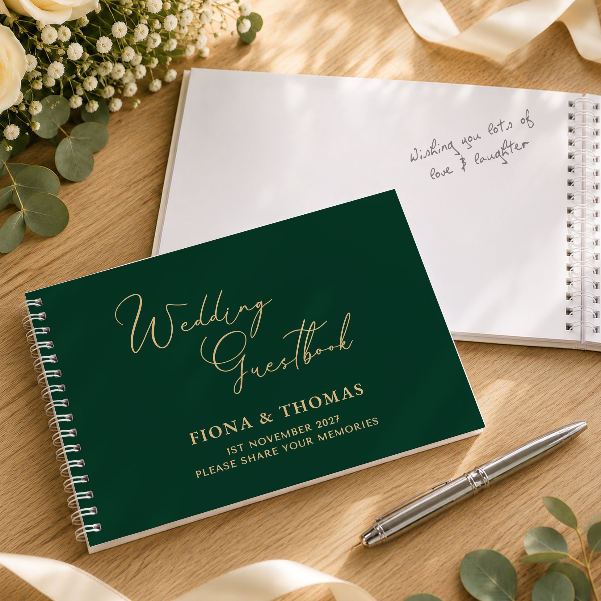 A personalised wedding guest book with a dark green cover and gold foil lettering, styled on a wooden table with a silver pen, white flowers, and eucalyptus leaves.