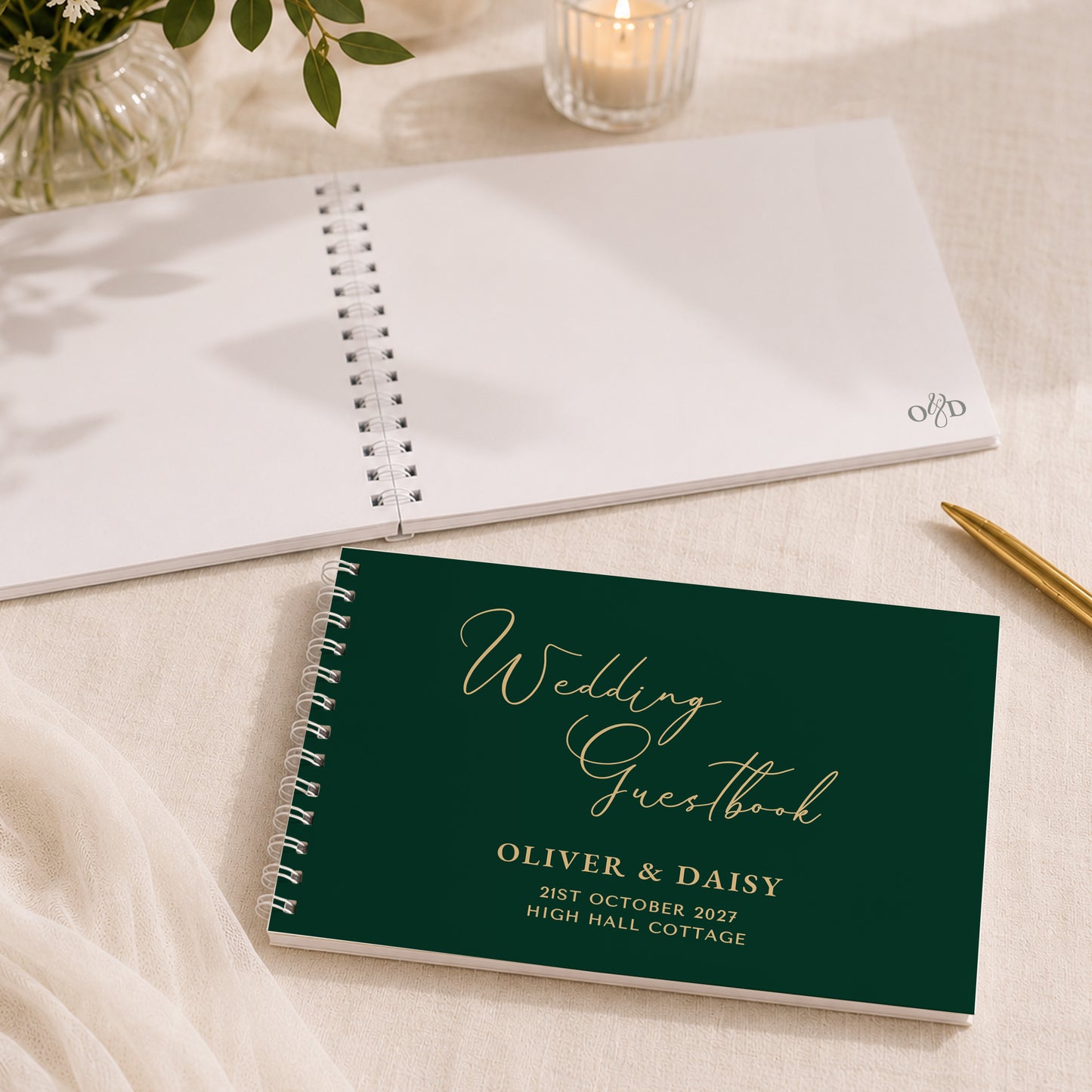 A personalised wedding guestbook with a dark green cover and gold foil lettering, shown in a styled flat-lay setting alongside an open white guestbook, a gold pen, and a lit candle.