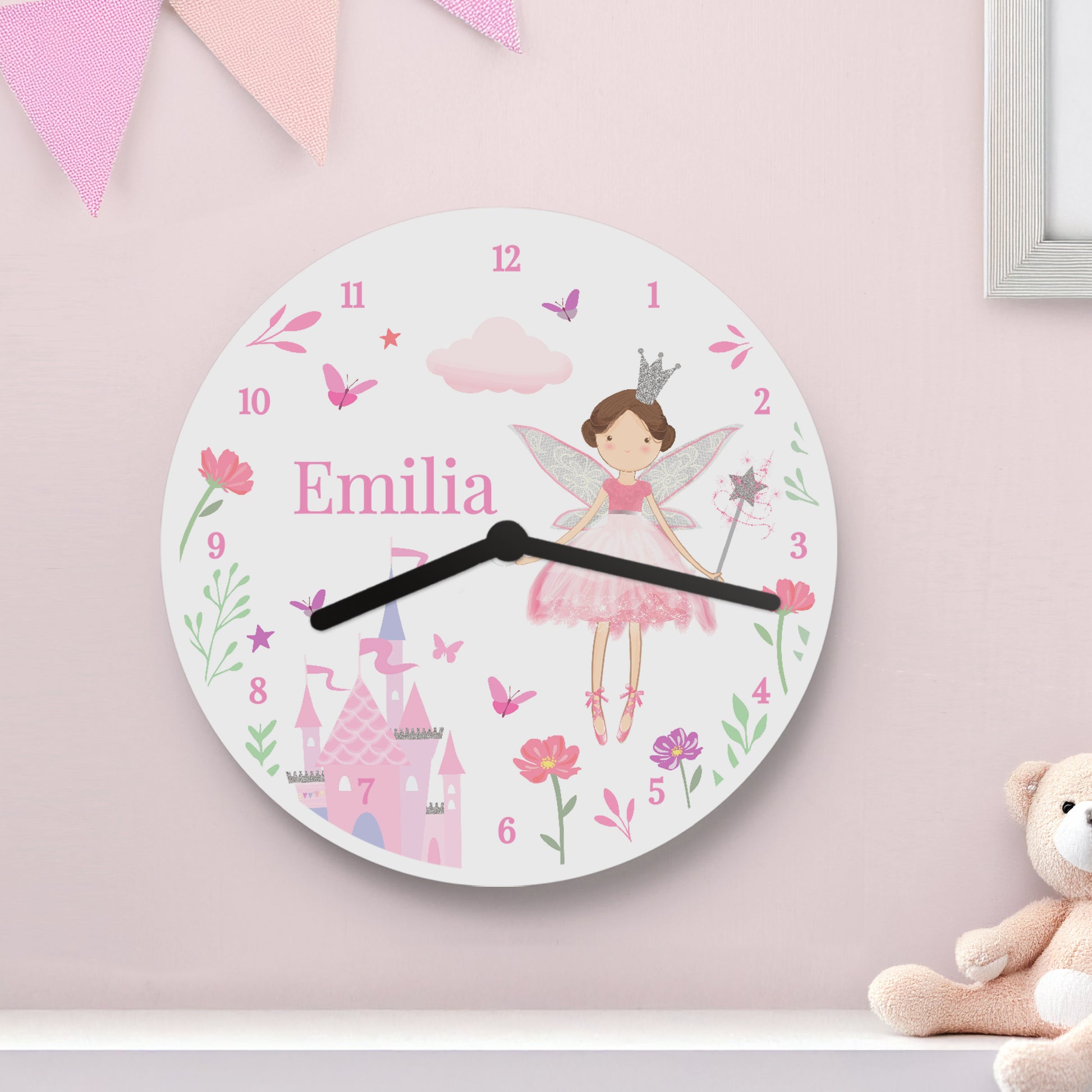 Children's wall clock with fairy design and 'Emilia' on a pink wall with a teddy bear.