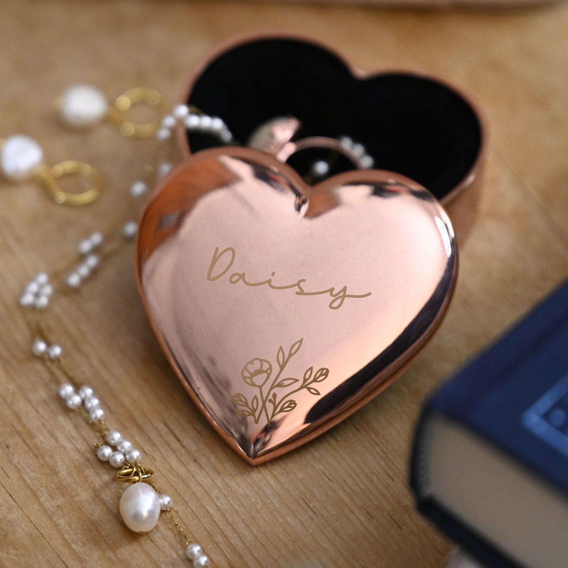 Heart-shaped rose gold trinket box with 'Daisy' engraving on a wooden surface with jewellery.