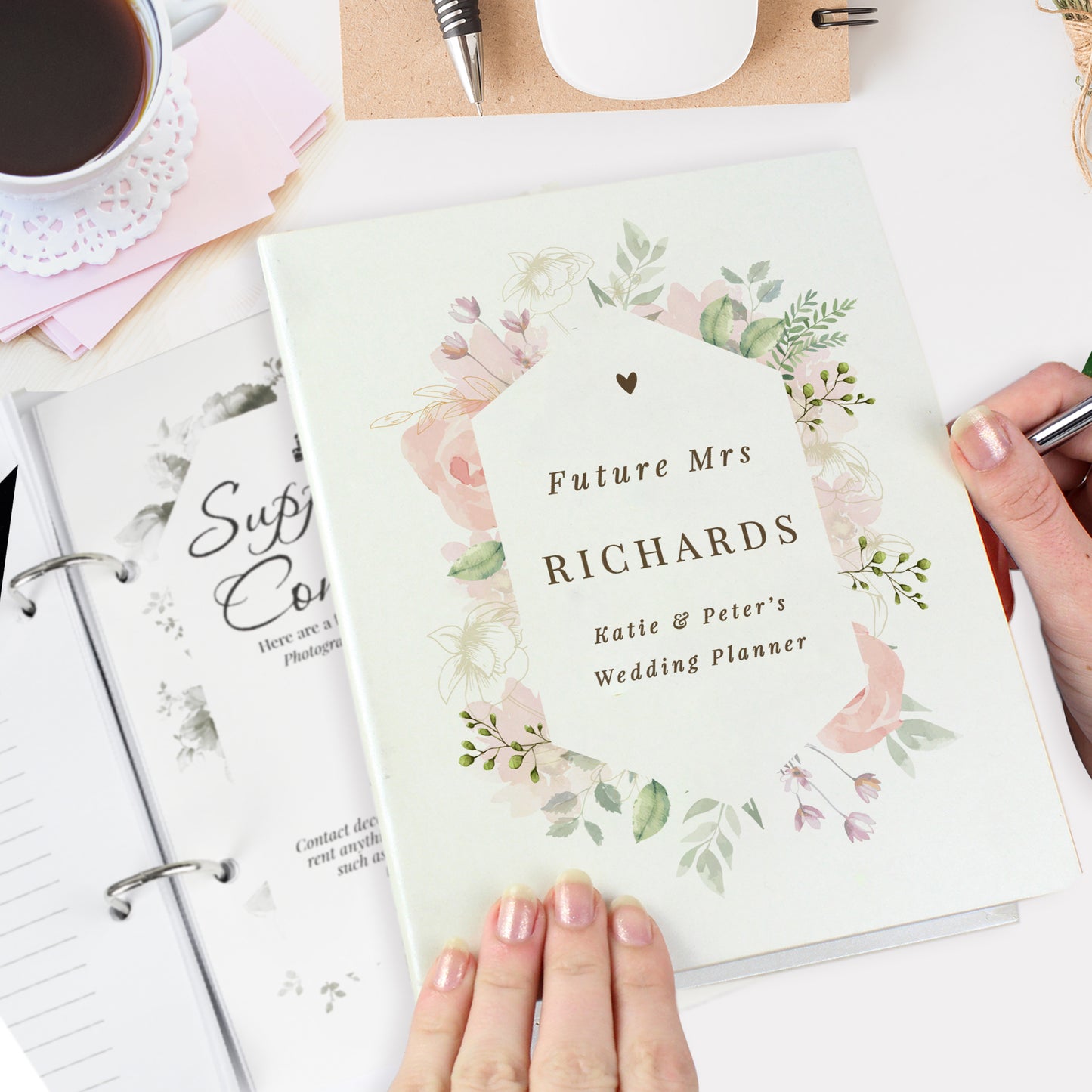 Hand holding a wedding planner book with floral design and text on a desk with coffee and stationery.