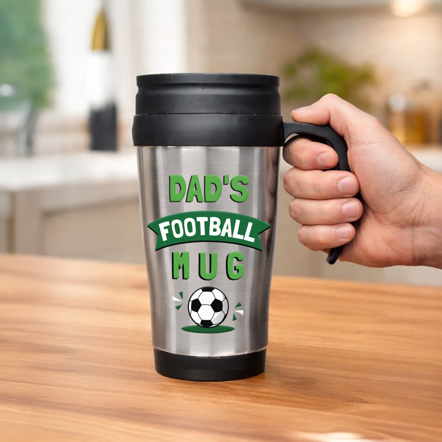 Silver travel mug with 'Dad's Football Mug' text and a hand holding it on a wooden surface.