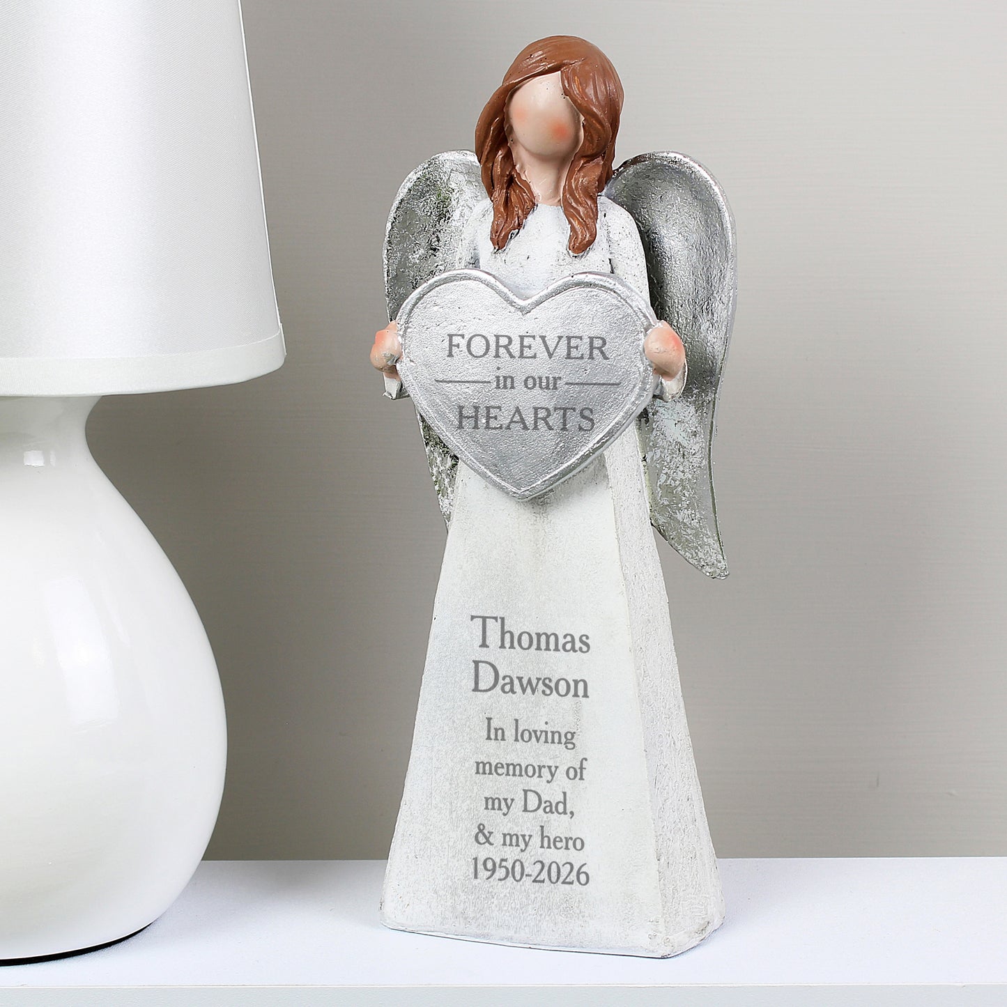 Angel figurine holding a heart-shaped plaque with engraved text on a white surface.