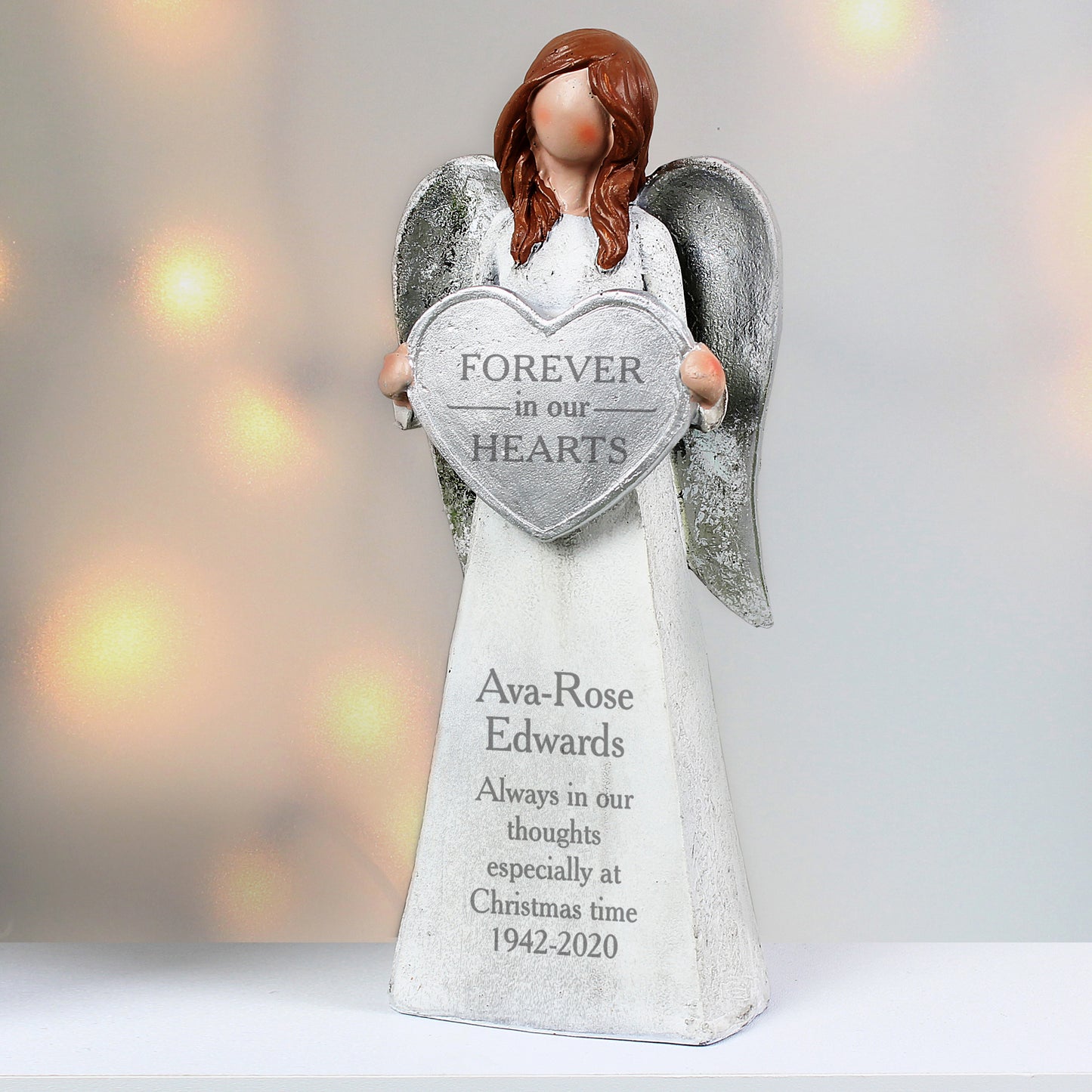 Angel figurine holding a heart-shaped 'Forever In Our Hearts' sign with personalised text on a blurred light background.