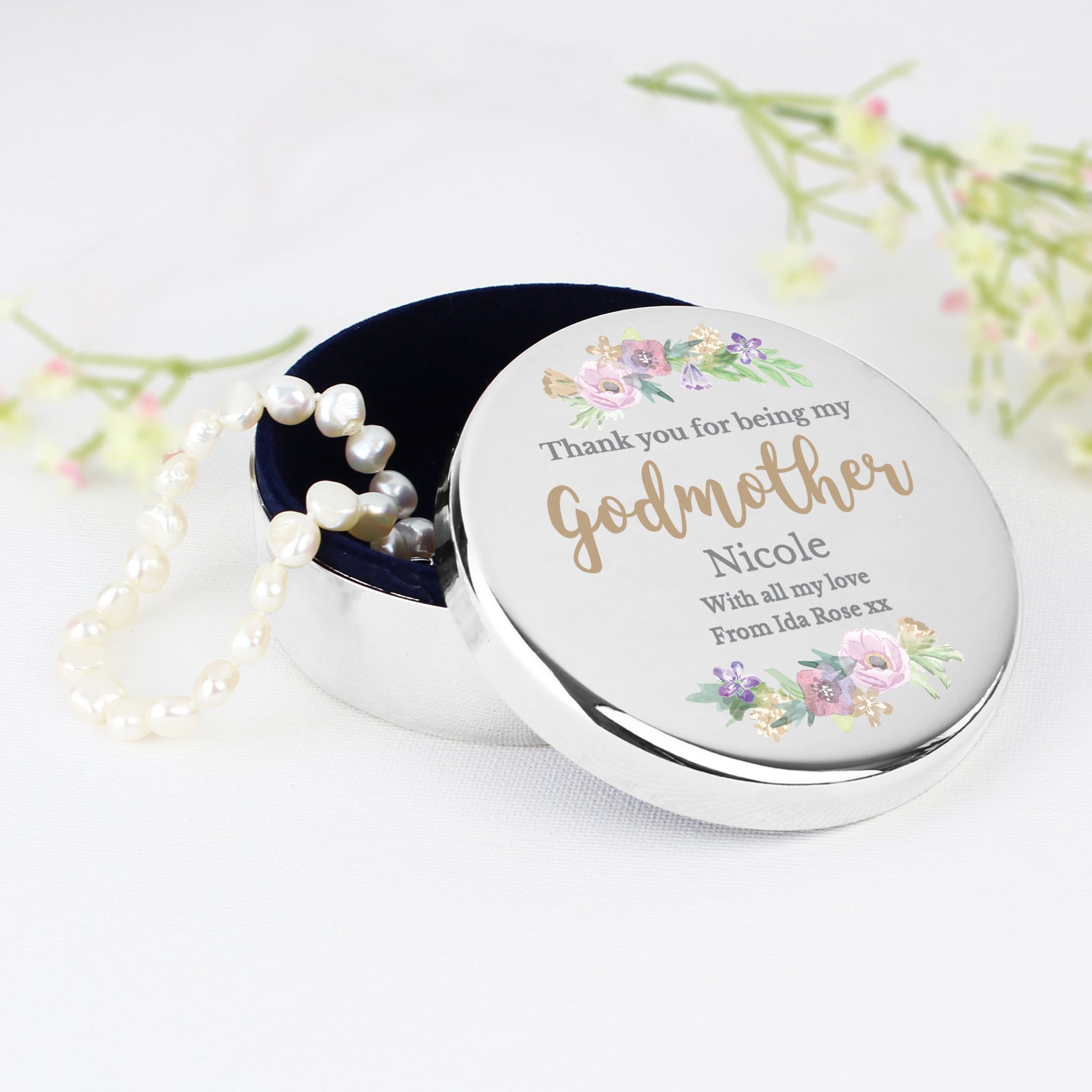 Round silver container with floral design and pearl necklace on a white background