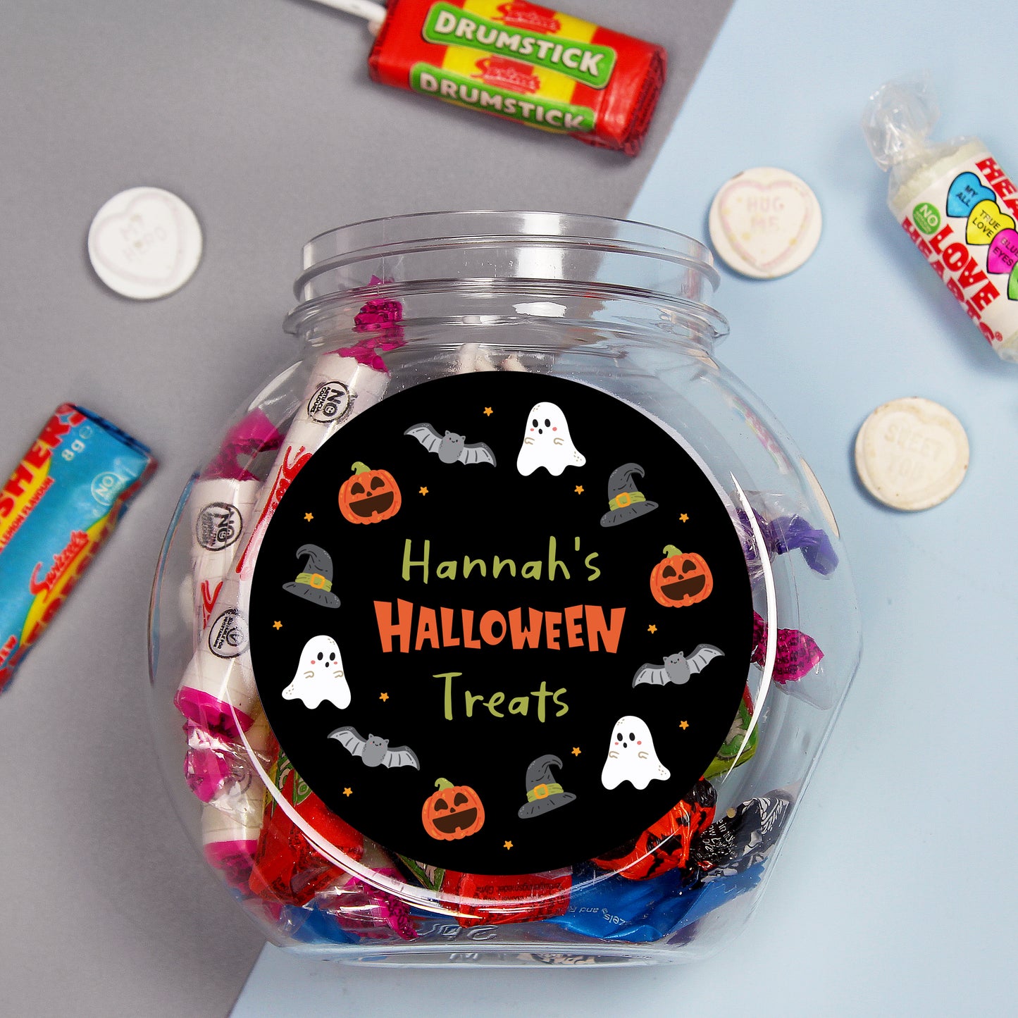 Clear candy jar with Halloween-themed label surrounded by assorted candies on a gray and blue background