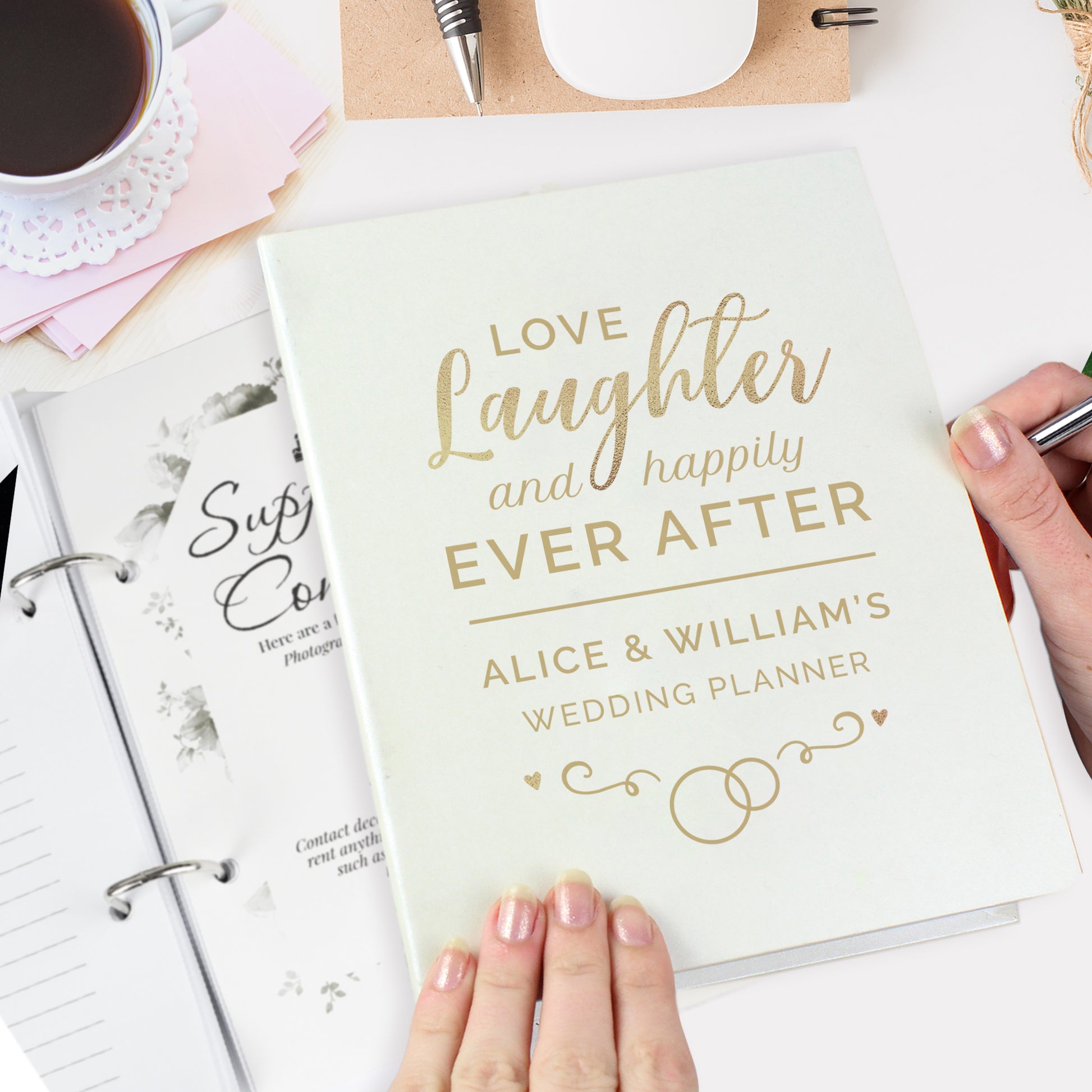 Wedding planner book titled 'Love, Laughter and Happily Ever After' held by a hand on a desk with coffee and stationery.