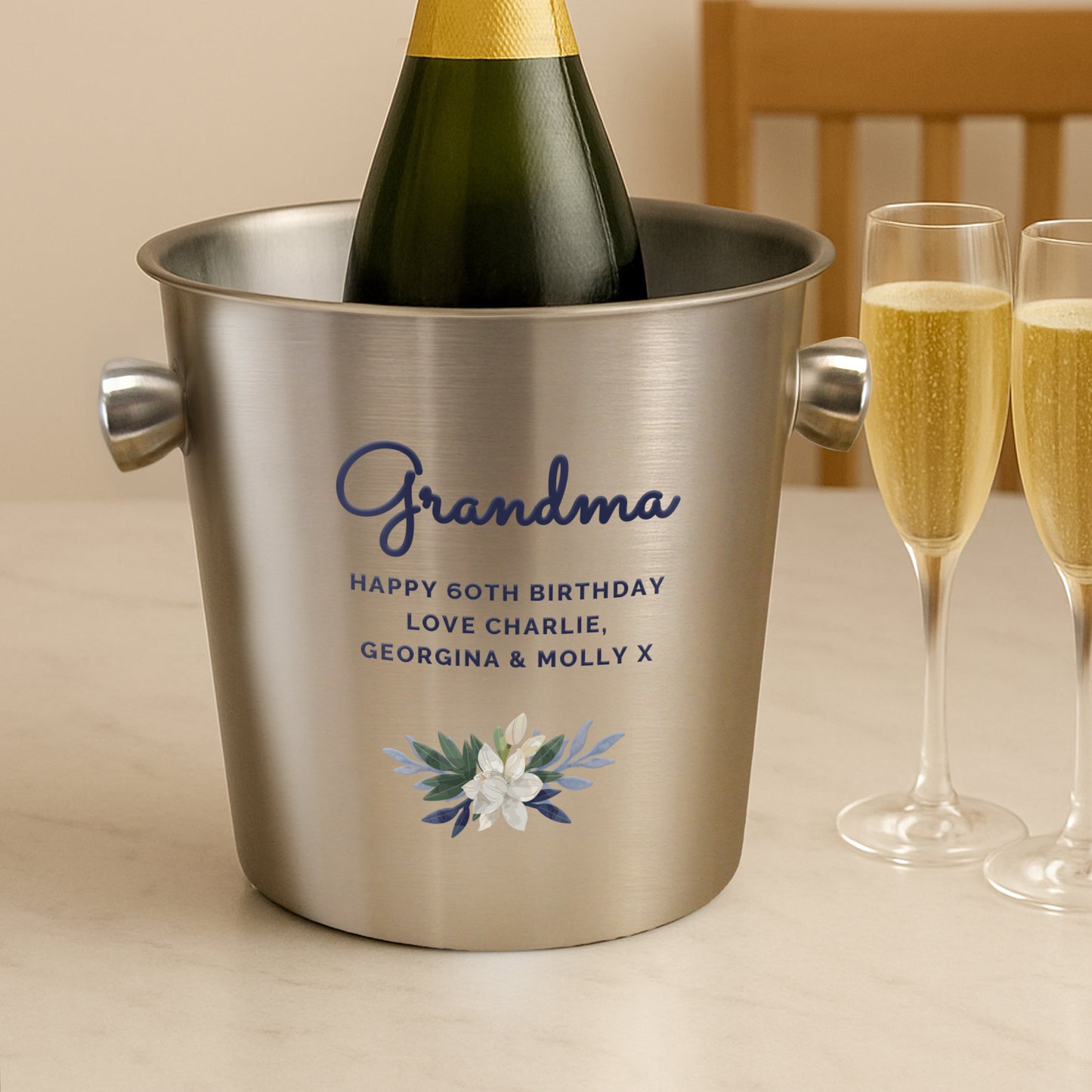 Silver ice bucket with 'Grandma' and birthday message, bottle of champagne inside, two glasses of champagne on a table. Champagne & Glasses not included