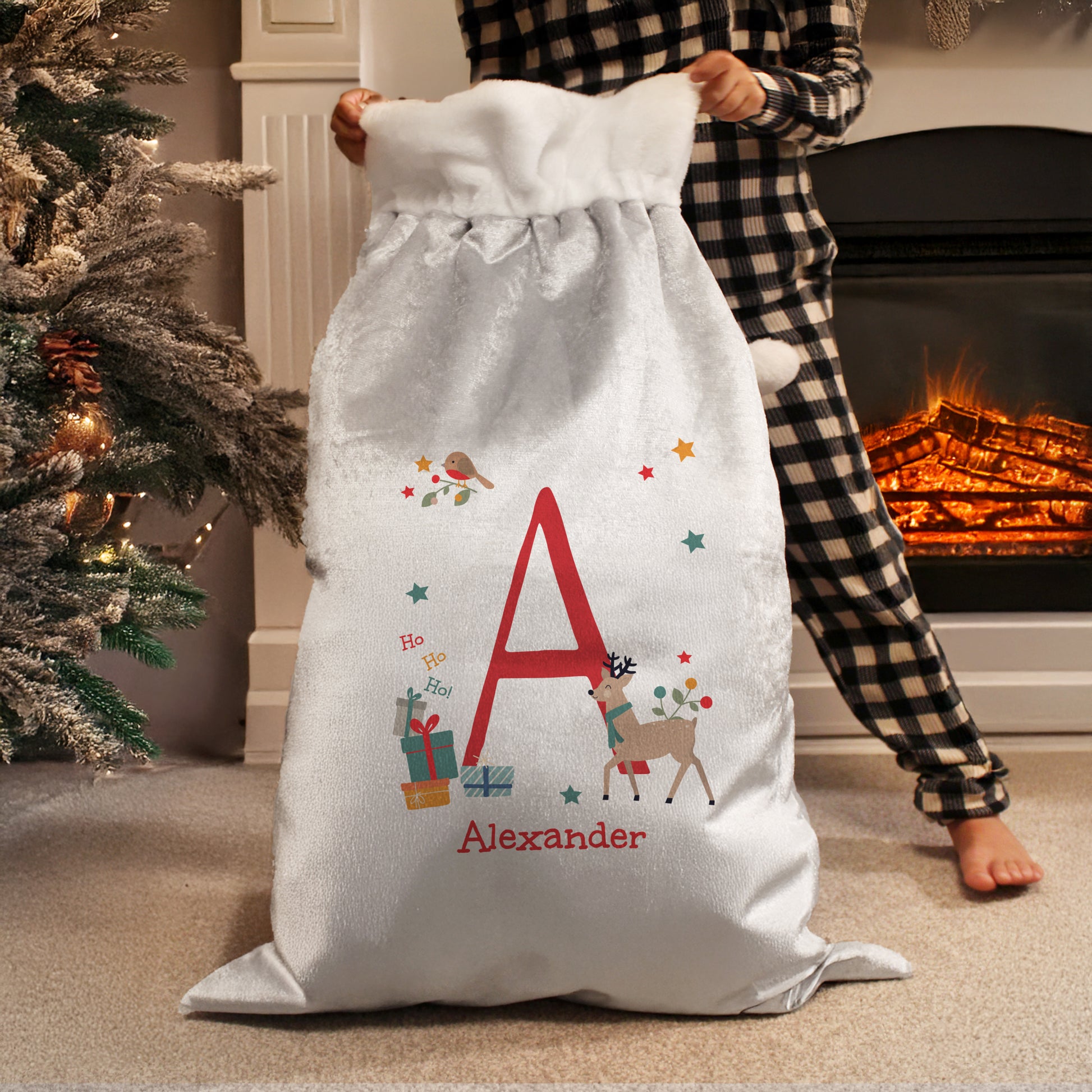Person holding a large white Christmas sack with decorative elements and the letter 'A' in front of a fireplace.