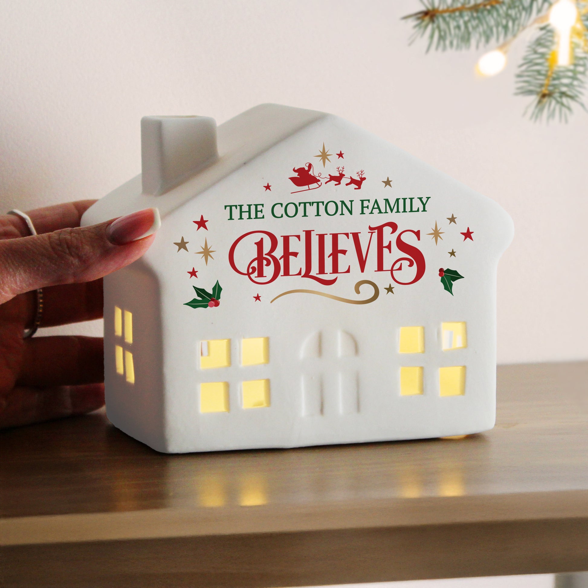 Someone holding a white ceramic Christmas LED house. Text reads The Cotton Family Believes.