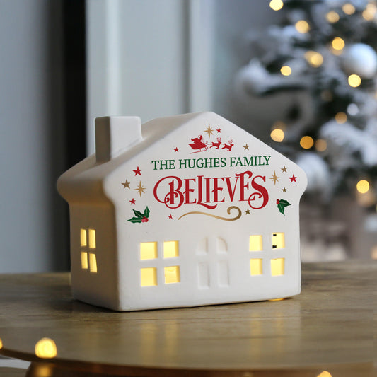 White ceramic Christmas LED house ornament on a wooden table with Christmas tree. Text reads The Hughes Family Believes.