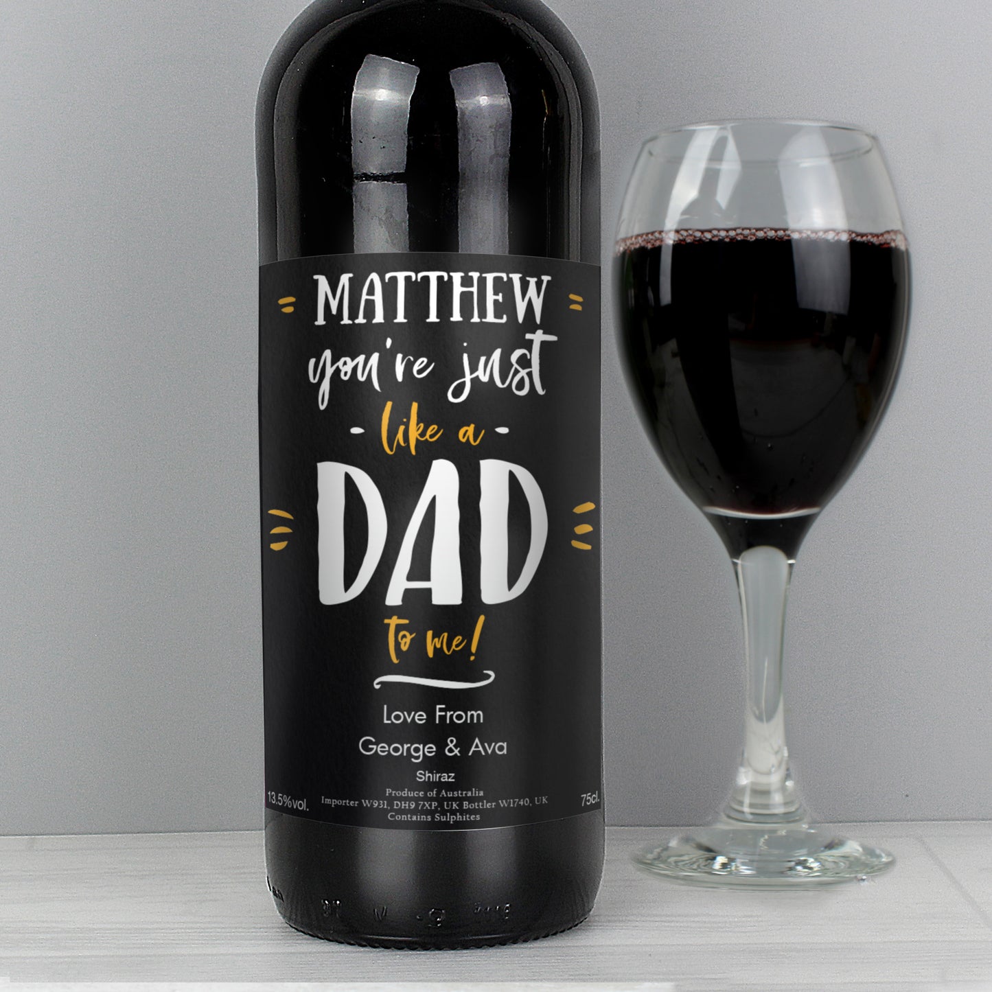 Wine bottle with a personalised label next to a glass of red wine on a grey background