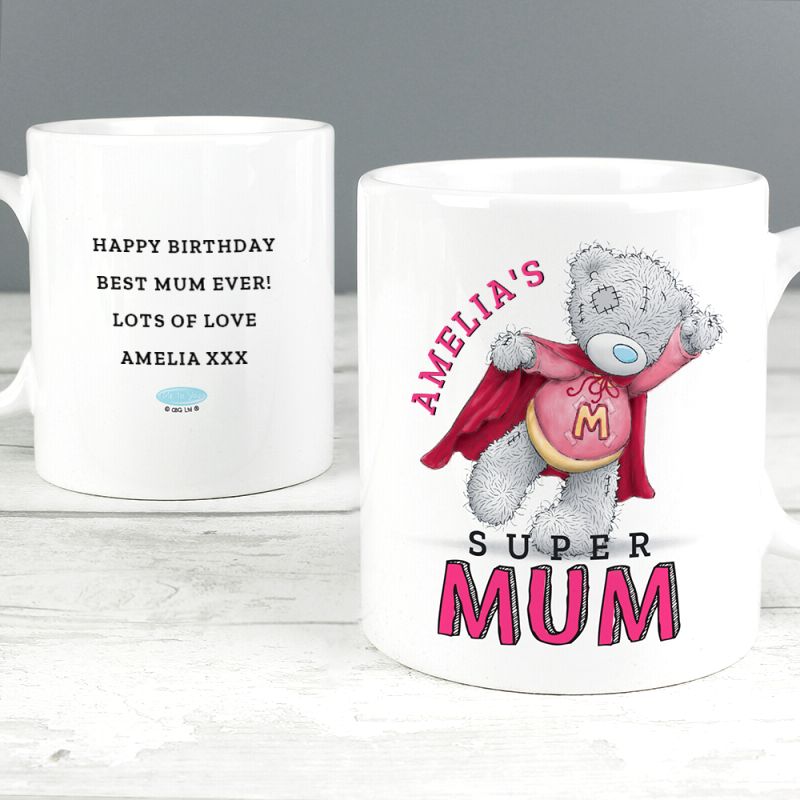 Two white ceramic mug images shown on a light wood surface against a grey background. The left mug has black personalised birthday text reading 'HAPPY BIRTHDAY BEST MUM EVER! LOTS OF LOVE AMELIA XXX' with a small MeToYou logo.