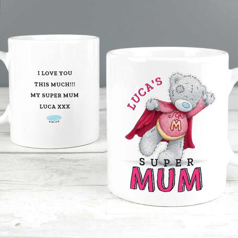 Two white ceramic mug images shown on a light wood surface against a grey background. The left mug has black text reading 'I LOVE YOU THIS MUCH!!! MY SUPER MUM LUCA XXX' with a small blue cloud graphic.