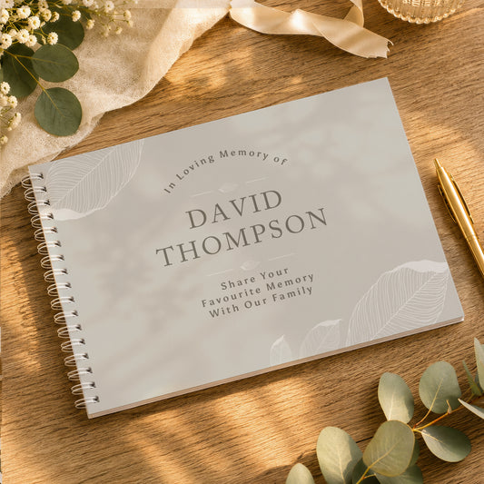A personalized spiral-bound memory book with a light grey cover, placed on a wooden tabletop with eucalyptus leaves, a gold pen, and soft fabric accents. The cover reads 'In Loving Memory of DAVID THOMPSON' and 'Share Your Favourite Memory With Our Family'.
