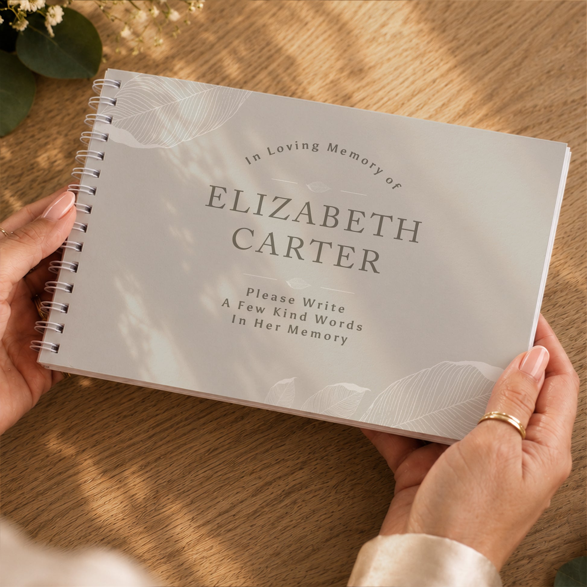A person holds a spiral-bound memorial guest book with a light grey cover printed with the text 'In Loving Memory of ELIZABETH CARTER' and 'Please Write A Few Kind Words In Her Memory.' The scene is photographed from above on a wooden surface with warm sunlight and shadows.