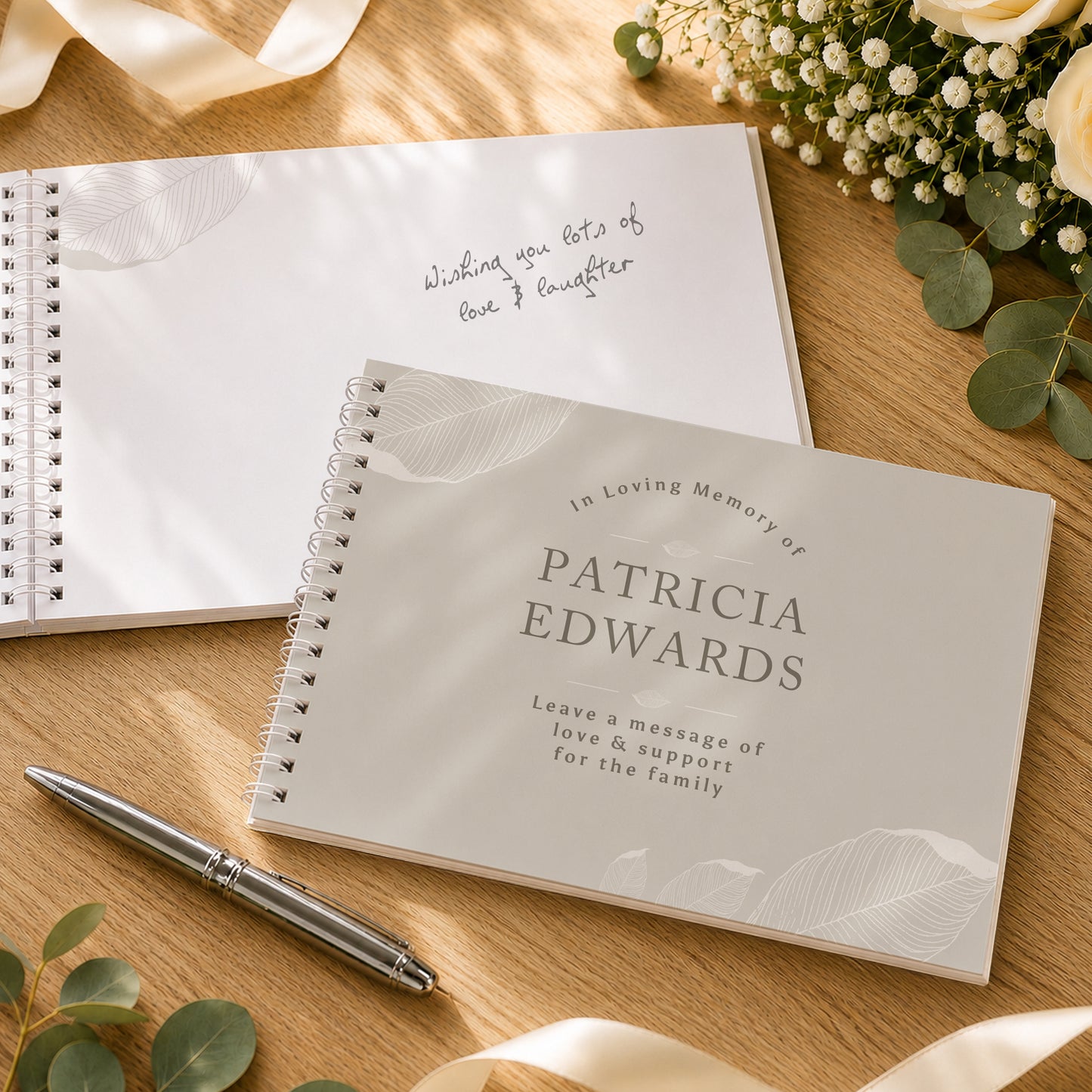 A spiral-bound memorial guest book in a light warm grey colour with a leaf pattern, shown closed with the text 'In Loving Memory of PATRICIA EDWARDS' and an open notebook behind it with handwritten text, styled on a wooden surface with a pen and flowers.
