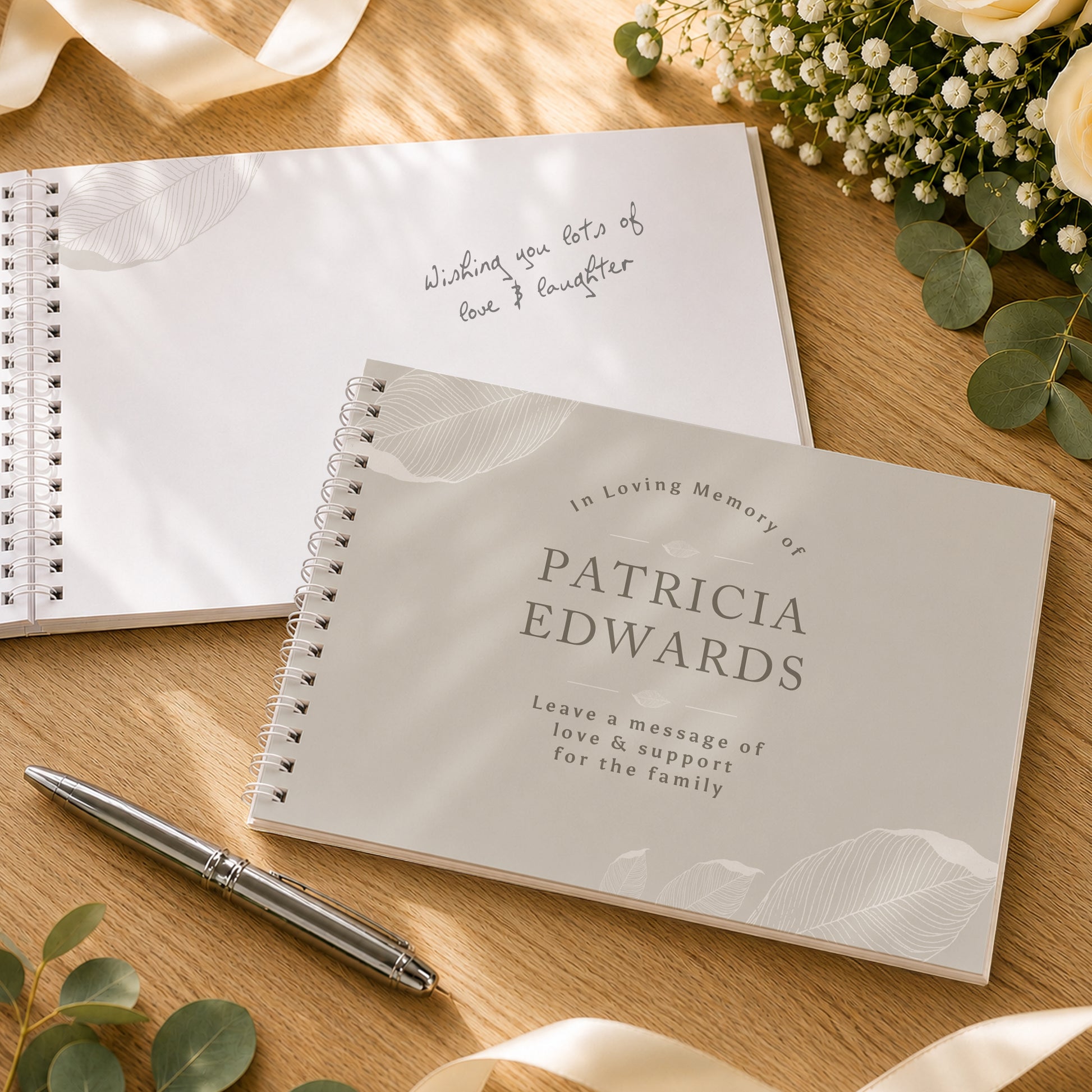 A spiral-bound memorial guest book in a light warm grey colour with a leaf pattern, shown closed with the text 'In Loving Memory of PATRICIA EDWARDS' and an open notebook behind it with handwritten text, styled on a wooden surface with a pen and flowers.