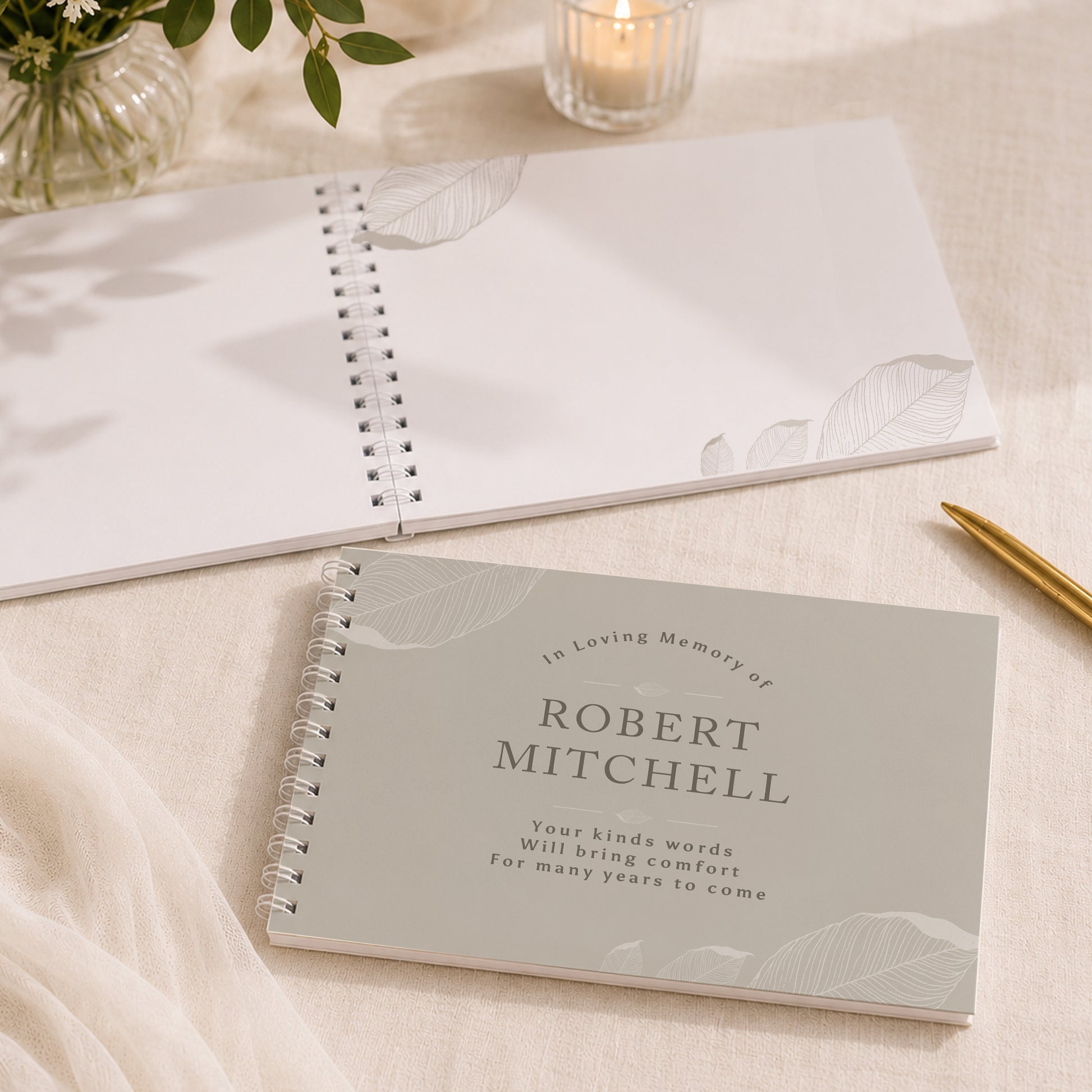 A personalised spiral-bound memorial notebook in a soft grey cover with the text 'In Loving Memory of ROBERT MITCHELL' and delicate leaf illustrations, styled on a cream fabric surface with a second open notebook, a candle, and a gold pen nearby.
