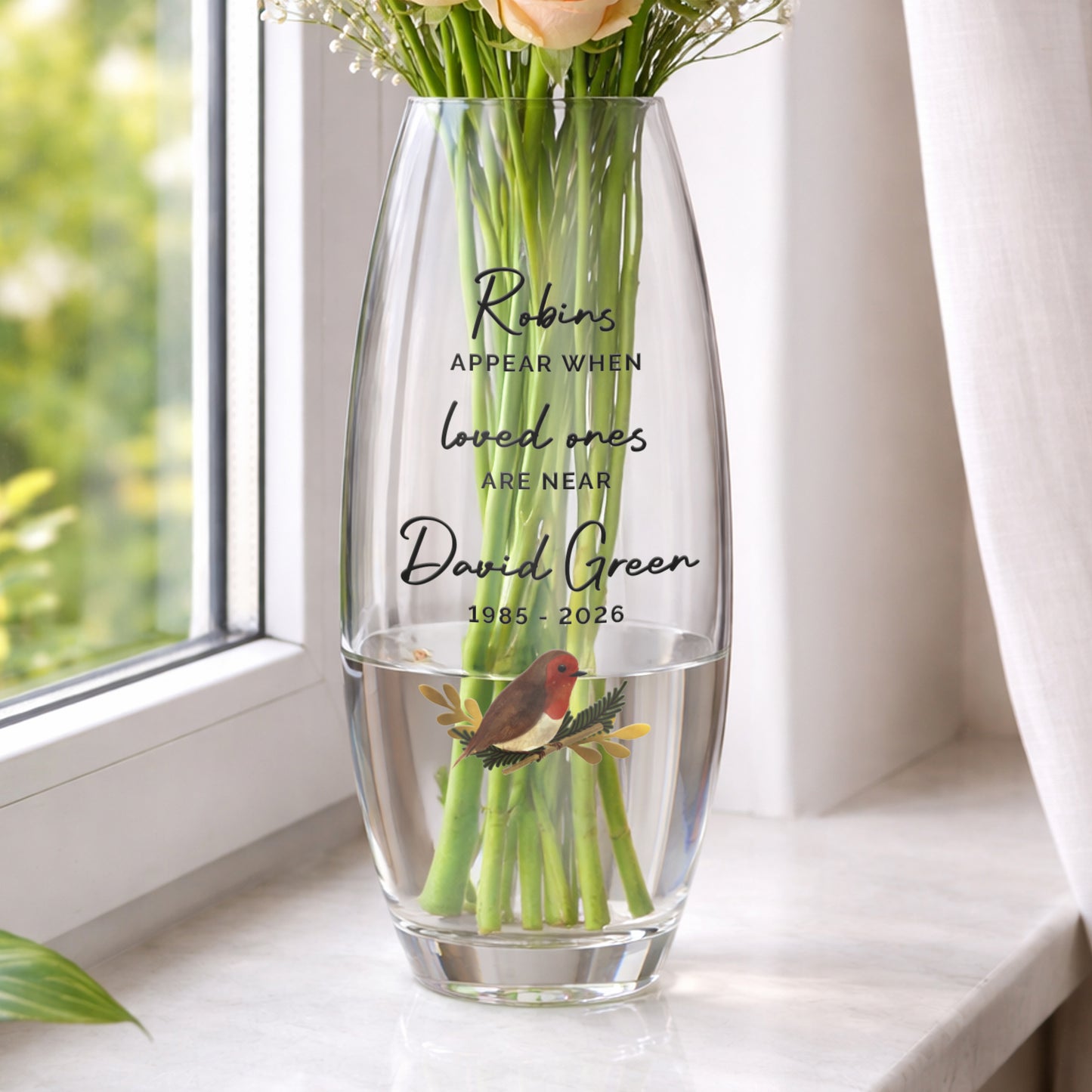 Clear glass vase with engraved text and a robin illustration, filled with water and flowers, on a windowsill.