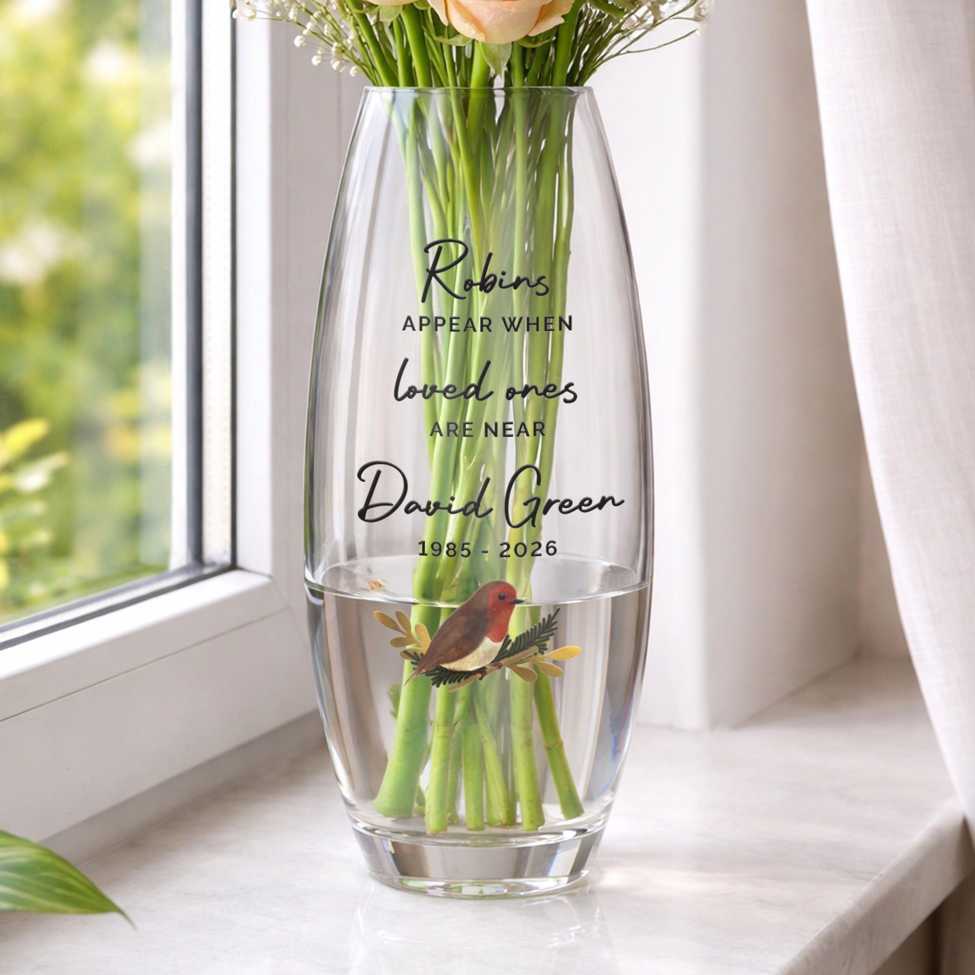 Clear glass vase with engraved text and a robin illustration, filled with water and flowers, on a windowsill.