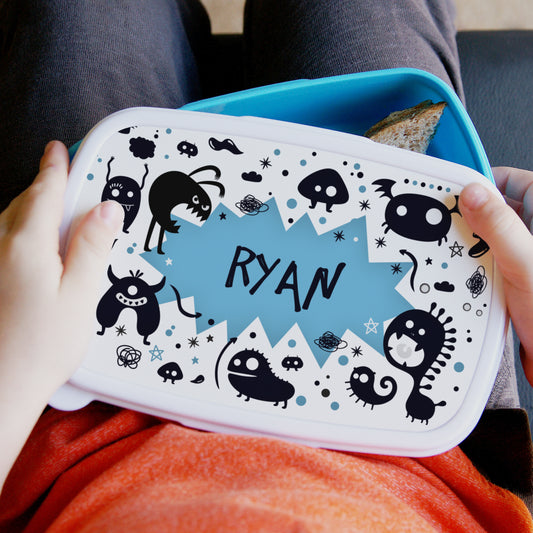 Children's lunchbox with cartoon monster design and 'Ryan' printed on it, held by a person.