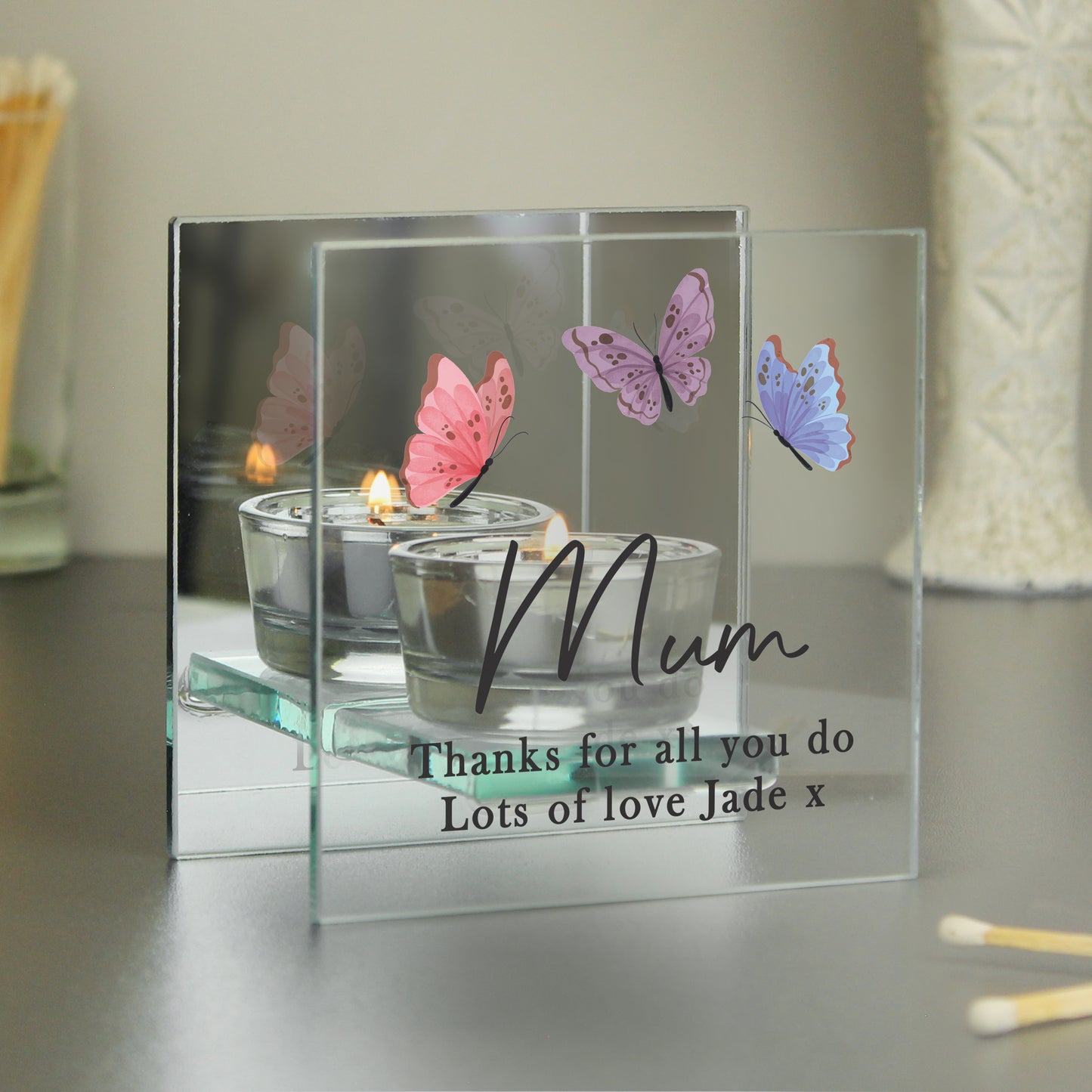 Glass candle holder with butterflies and a personalised message on a reflective surface.