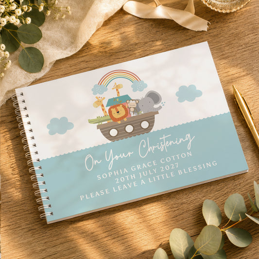 A personalized spiral-bound baby christening guest book with a Noah's Ark illustration, displayed on a wooden tabletop with decorative eucalyptus leaves, a gold pen, and a cream ribbon.