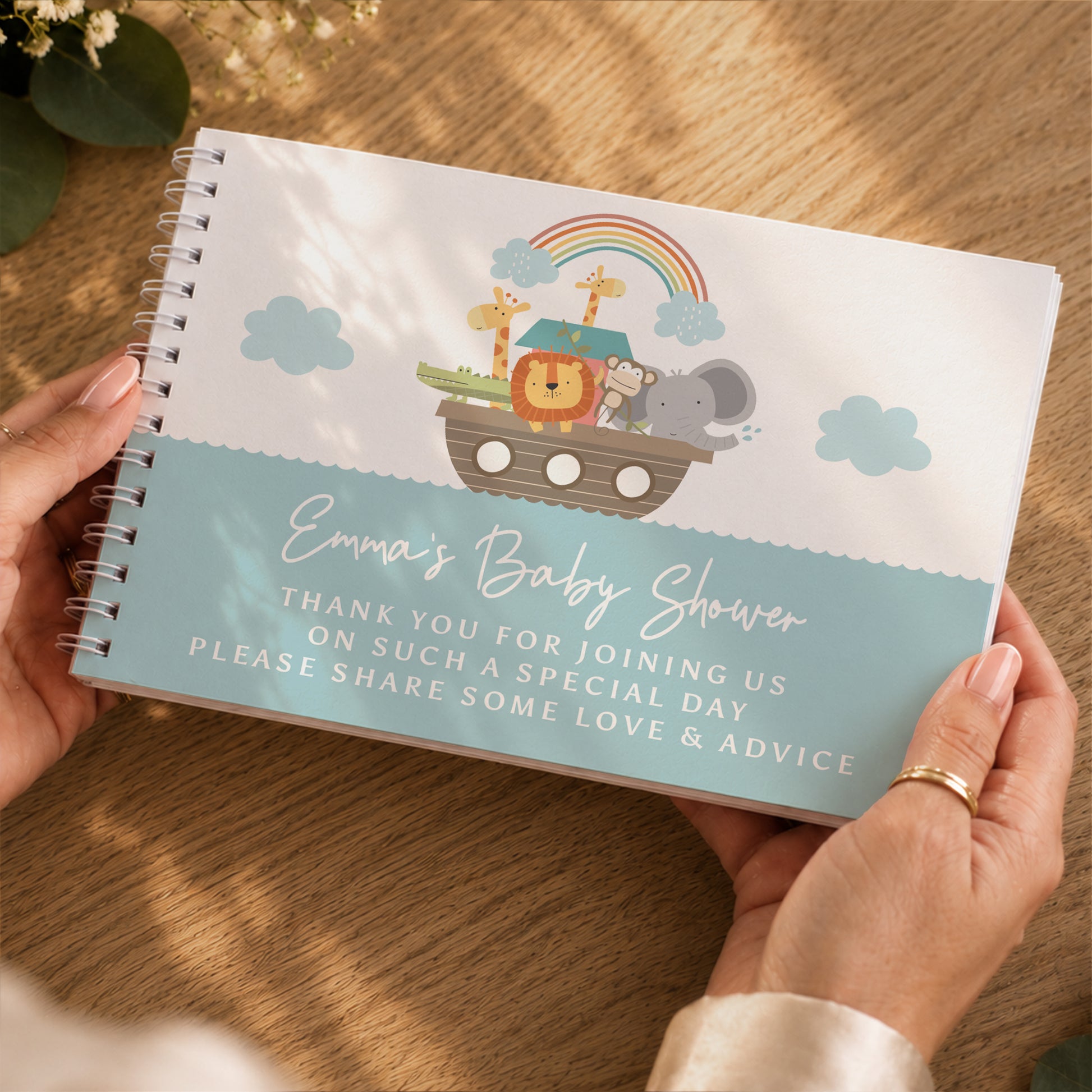 A person holds a spiral-bound baby shower notebook over a wooden table. The notebook has a white top half with a cartoon Noah's Ark illustration and a light blue bottom half with the text 'Emma's Baby Shower' and a thank-you message.