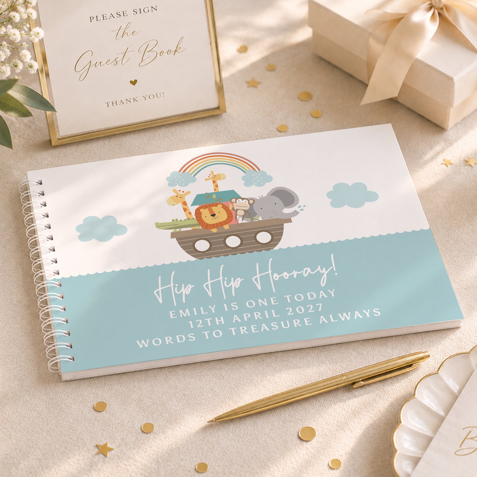 A spiral-bound guest book with a Noah's Ark illustration and the text 'Hip Hip Hooray! Emily is one today 12th April 2027 Words to Treasure Always' is styled on a beige fabric surface with gold star confetti, a gold pen, a framed guest book sign, and a gift box.
