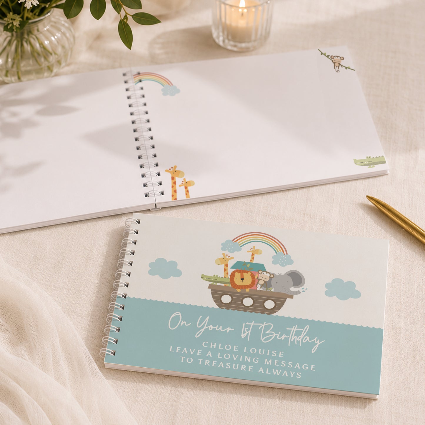 A spiral-bound children's keepsake notebook lies open on a light fabric surface, with a matching closed notebook in front showing a Noah's Ark illustration and the text 'On Your 1st Birthday' plus a personalized message. Decorative props including a candle, plant leaves, and a gold pen are visible around it.