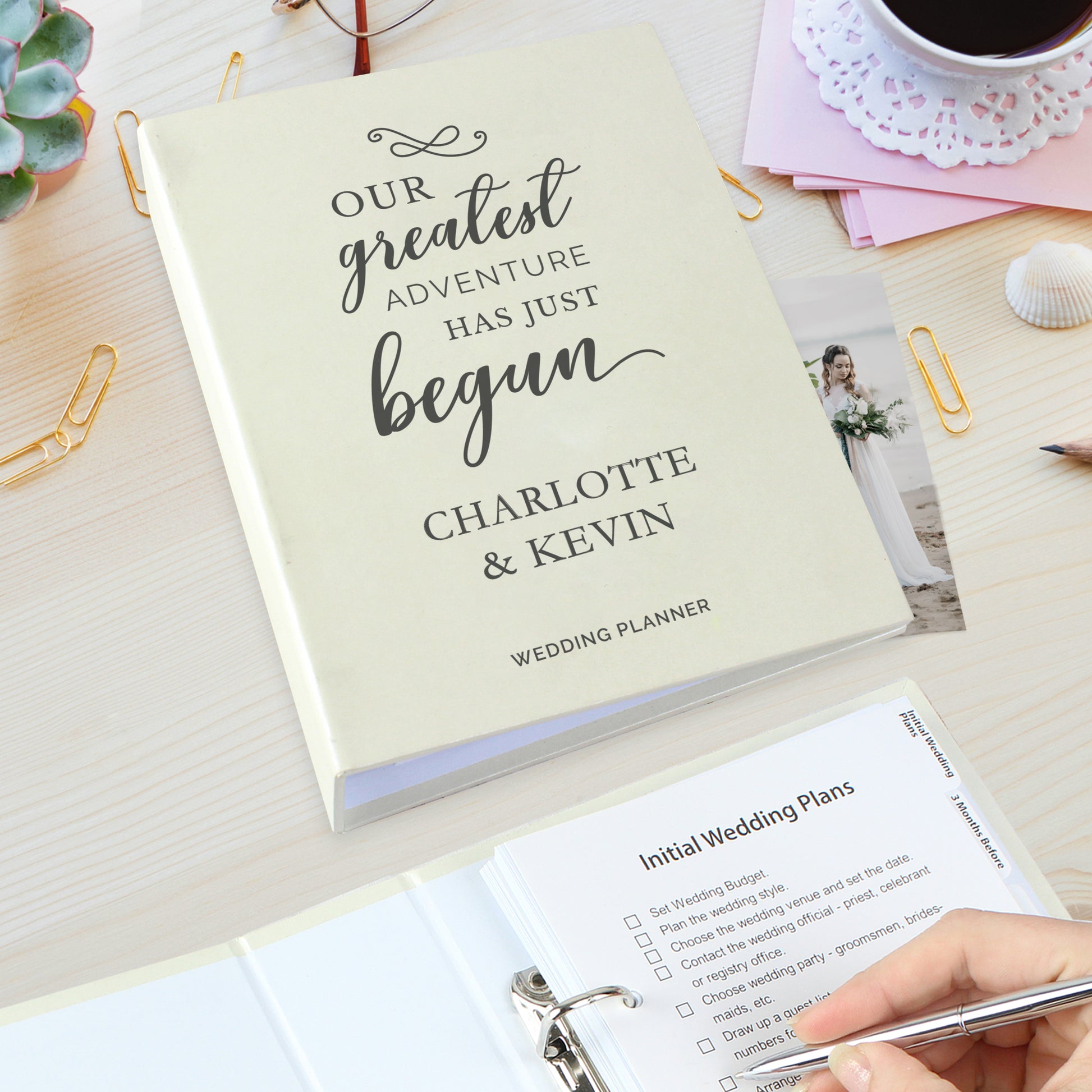 Wedding planner book with 'Our greatest adventure has just begun' text, surrounded by stationery items on a wooden surface.