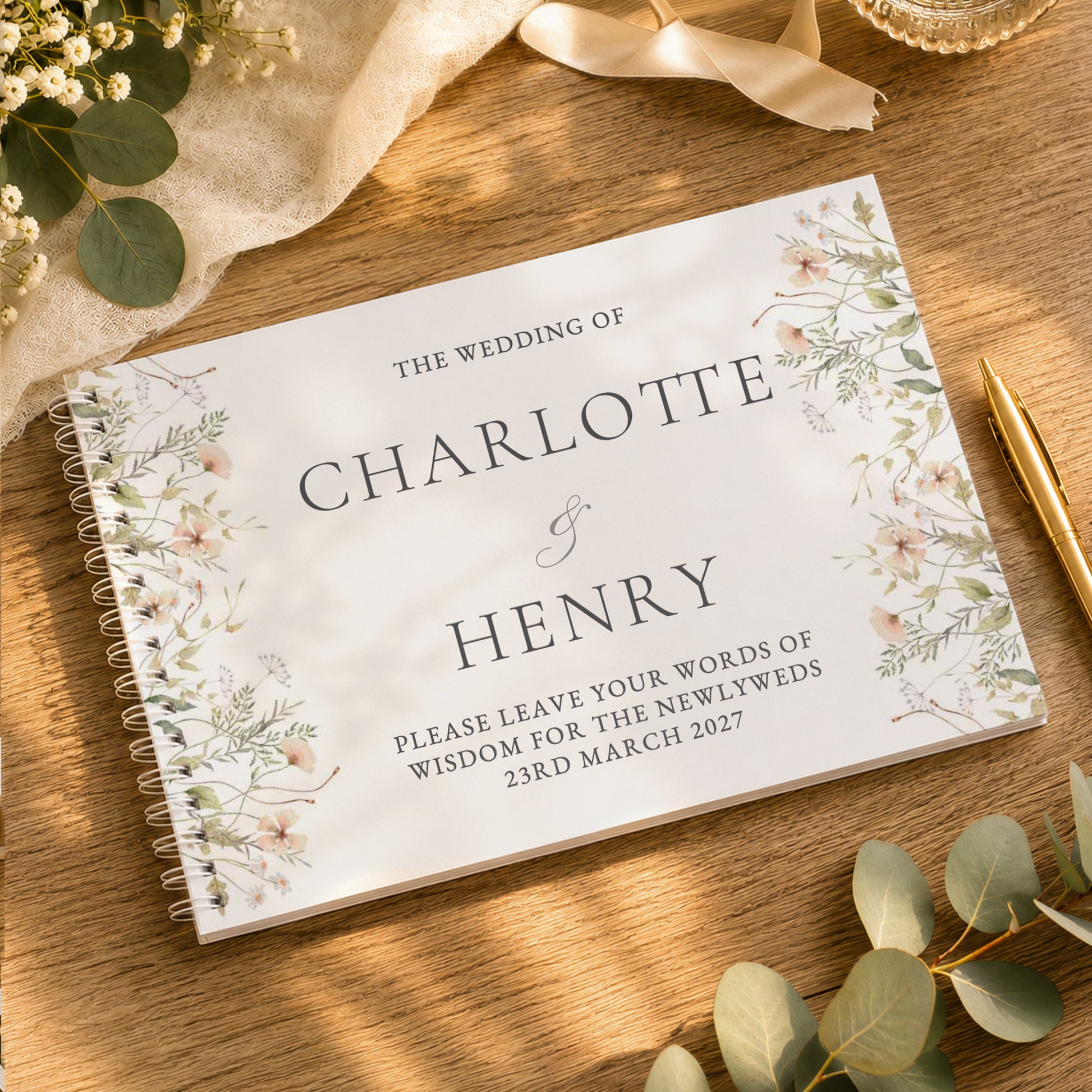 A personalised spiral-bound wedding guest book with floral illustrations on a wooden table, styled with eucalyptus leaves, a gold pen, and a cream ribbon.