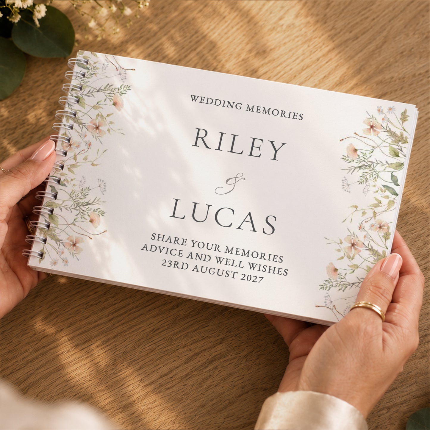 A person holds a personalised spiral-bound wedding memory book with floral illustrations on a wooden table. The book cover reads 'WEDDING MEMORIES', 'RILEY & LUCAS', and 'SHARE YOUR MEMORIES ADVICE AND WELL WISHES 23RD AUGUST 2027'.