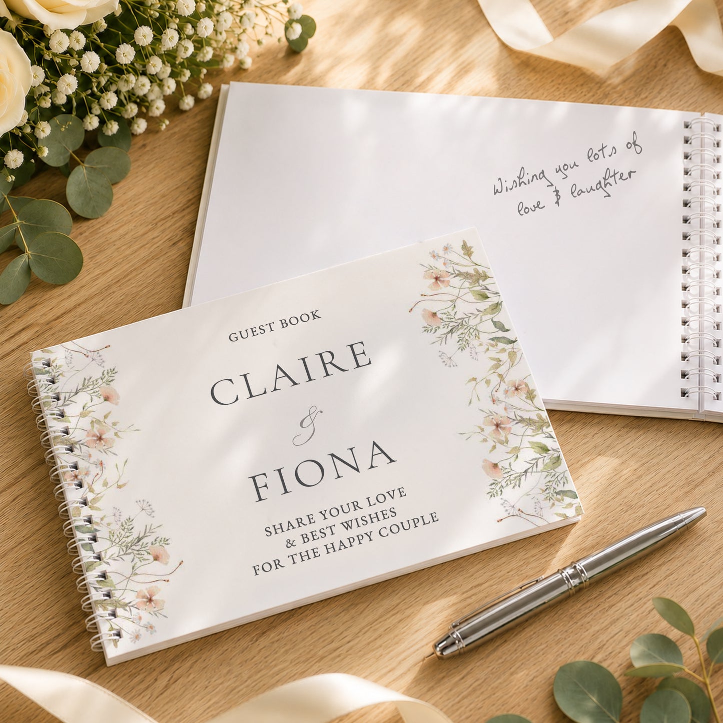 A personalised spiral guest book with floral illustrations on the cover, placed on a wooden surface with a silver pen, additional guest books, and floral props.