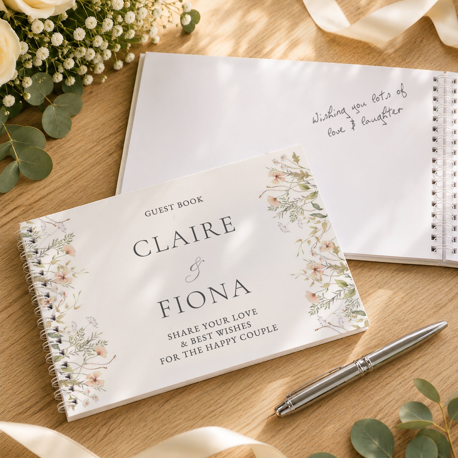 A personalised spiral guest book with floral illustrations on the cover, placed on a wooden surface with a silver pen, additional guest books, and floral props.