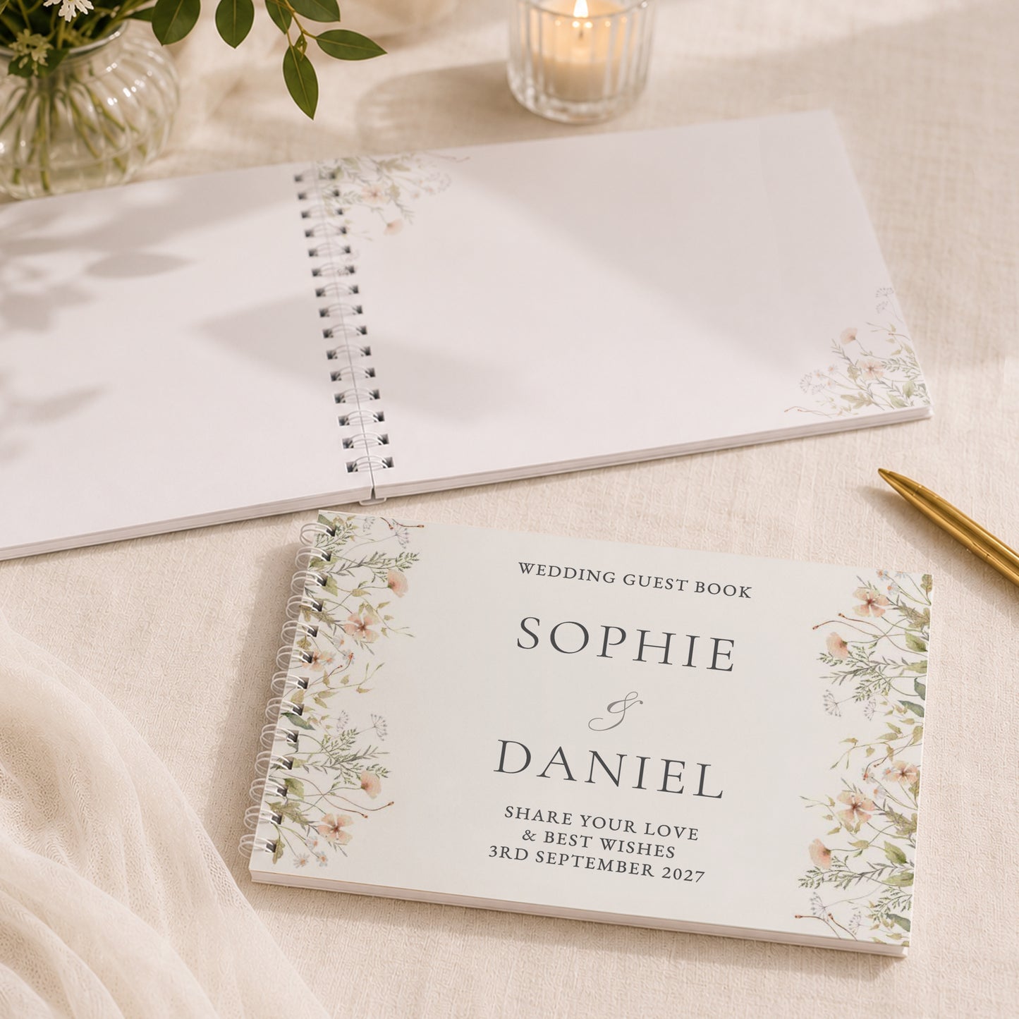A personalised wedding guest book with floral watercolor corner designs and elegant typography, displayed on a beige fabric surface alongside a blank copy of the same book, a lit candle, and a gold pen.