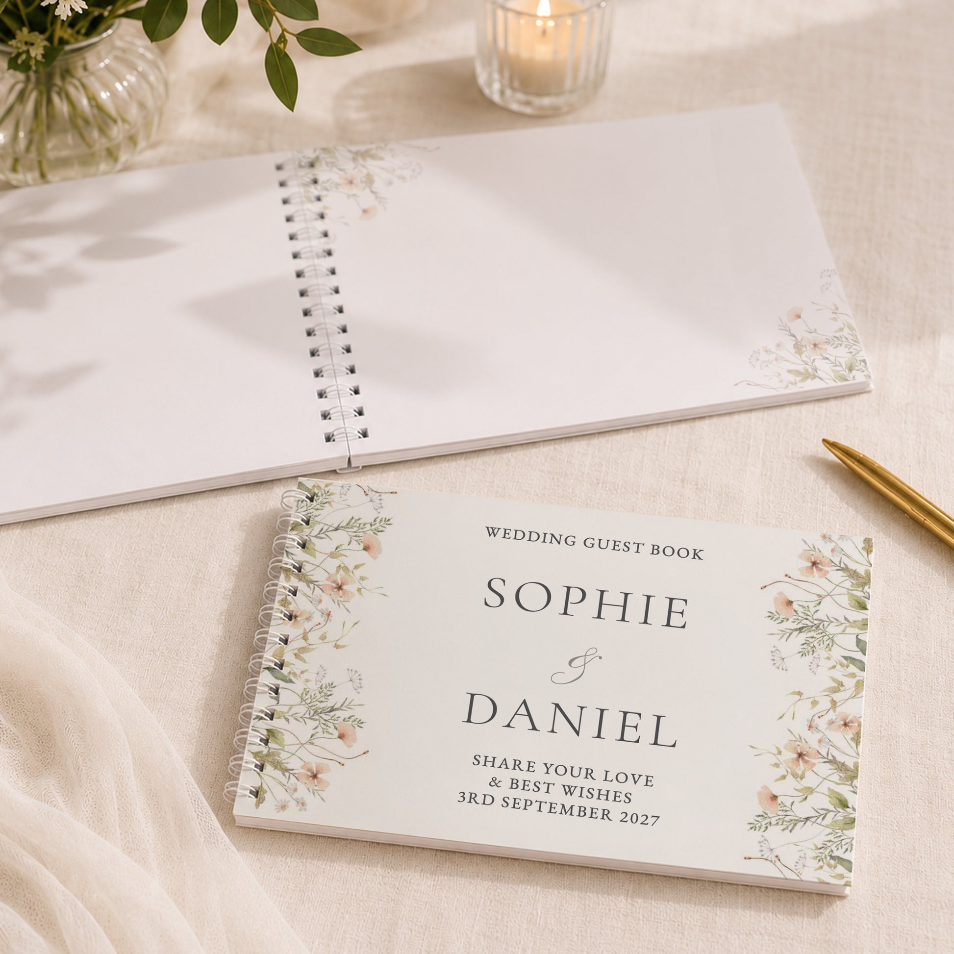 A personalised wedding guest book with floral watercolor corner designs and elegant typography, displayed on a beige fabric surface alongside a blank copy of the same book, a lit candle, and a gold pen.