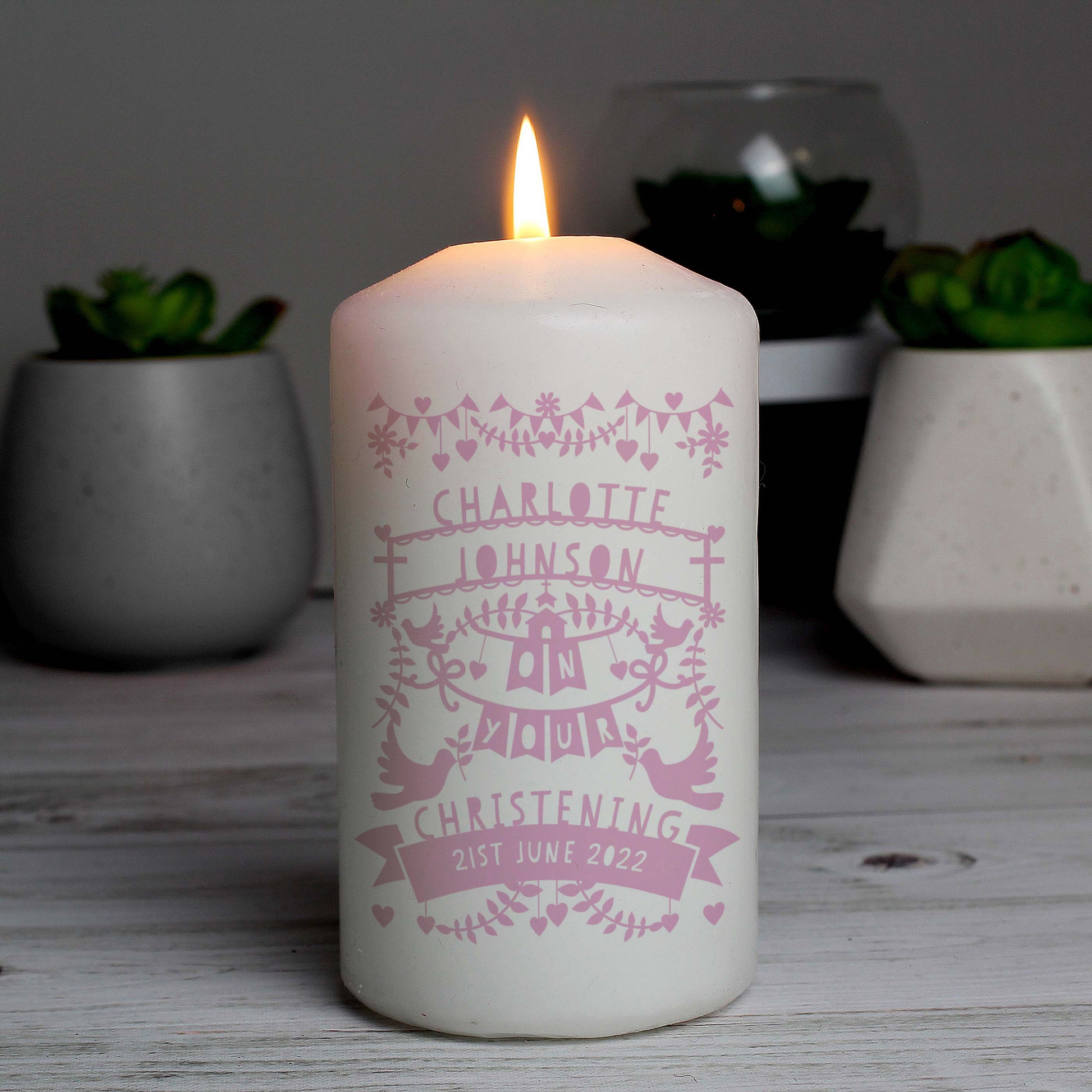 White candle with pink text and design, lit on a wooden surface with plants in the background